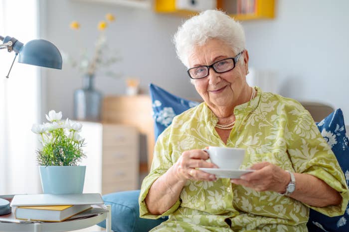 Elderly woman with glasses smiling, holding a teacup, seated indoors with books and a plant nearby