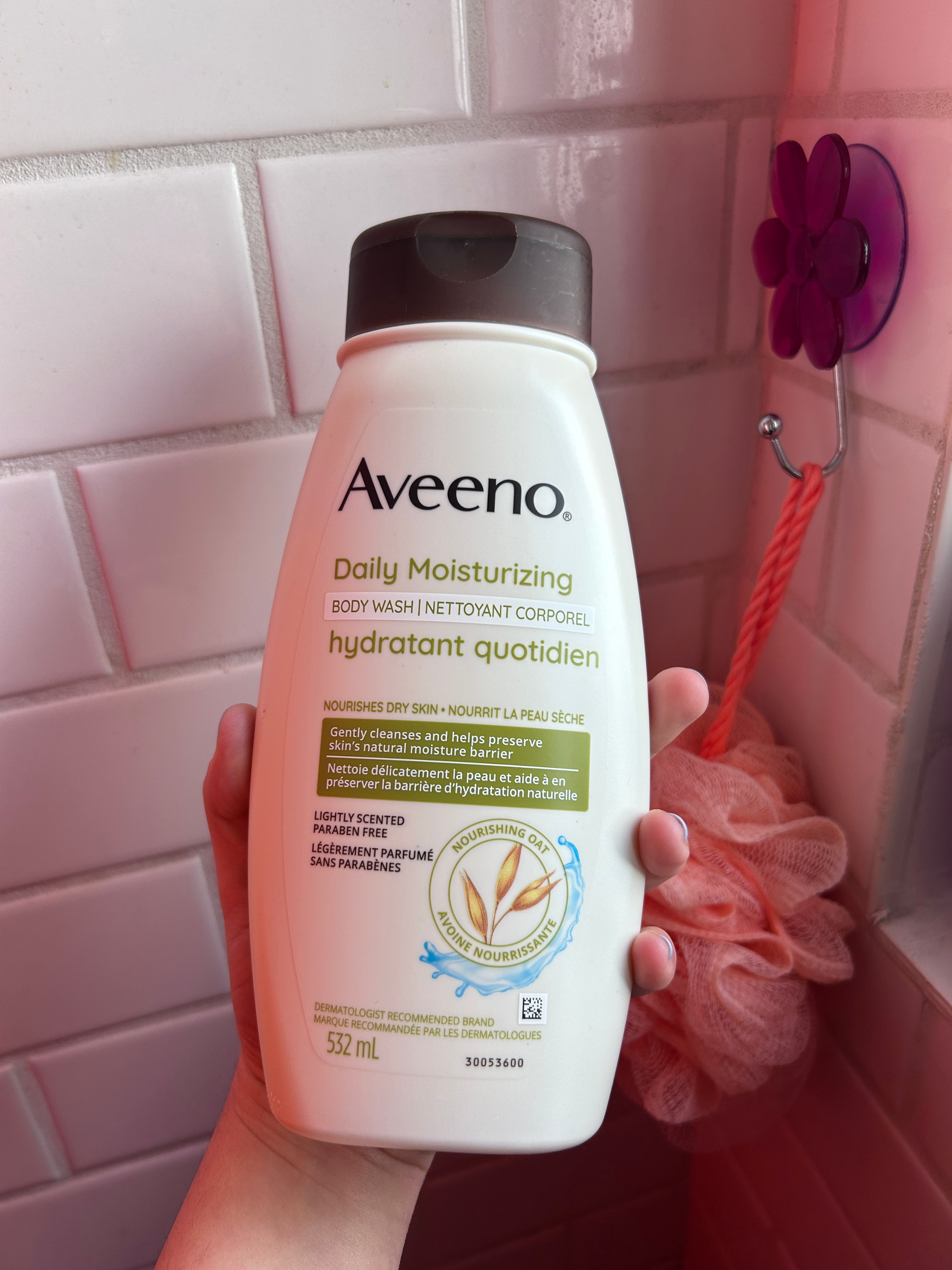 Hand holding a bottle of Aveeno Daily Moisturizing Body Wash next to a shower loofah