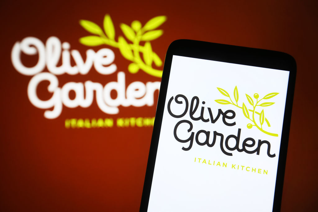 Smartphone displaying Olive Garden logo with a blurred restaurant sign in the background
