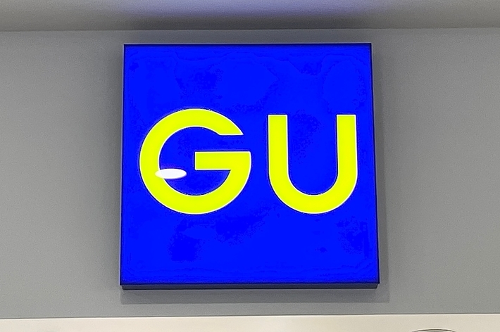 Sign with the letters &ldquo;GU&rdquo; against a blue background, mounted on a wall with ceiling lights visible
