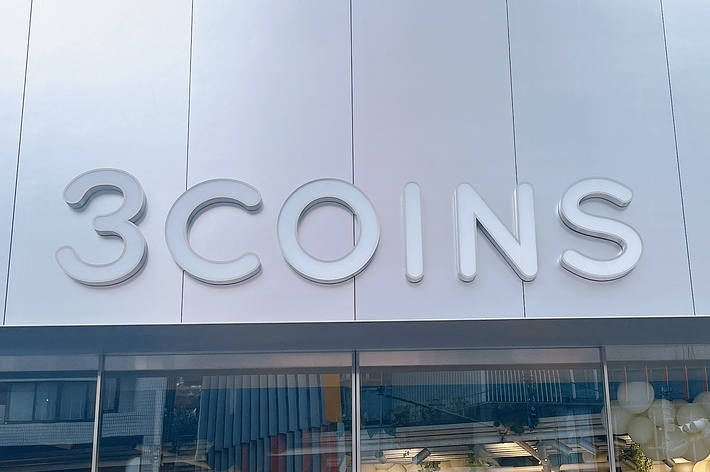 店舗の看板「3COINS」ロゴと下に掲示された広告が映る。