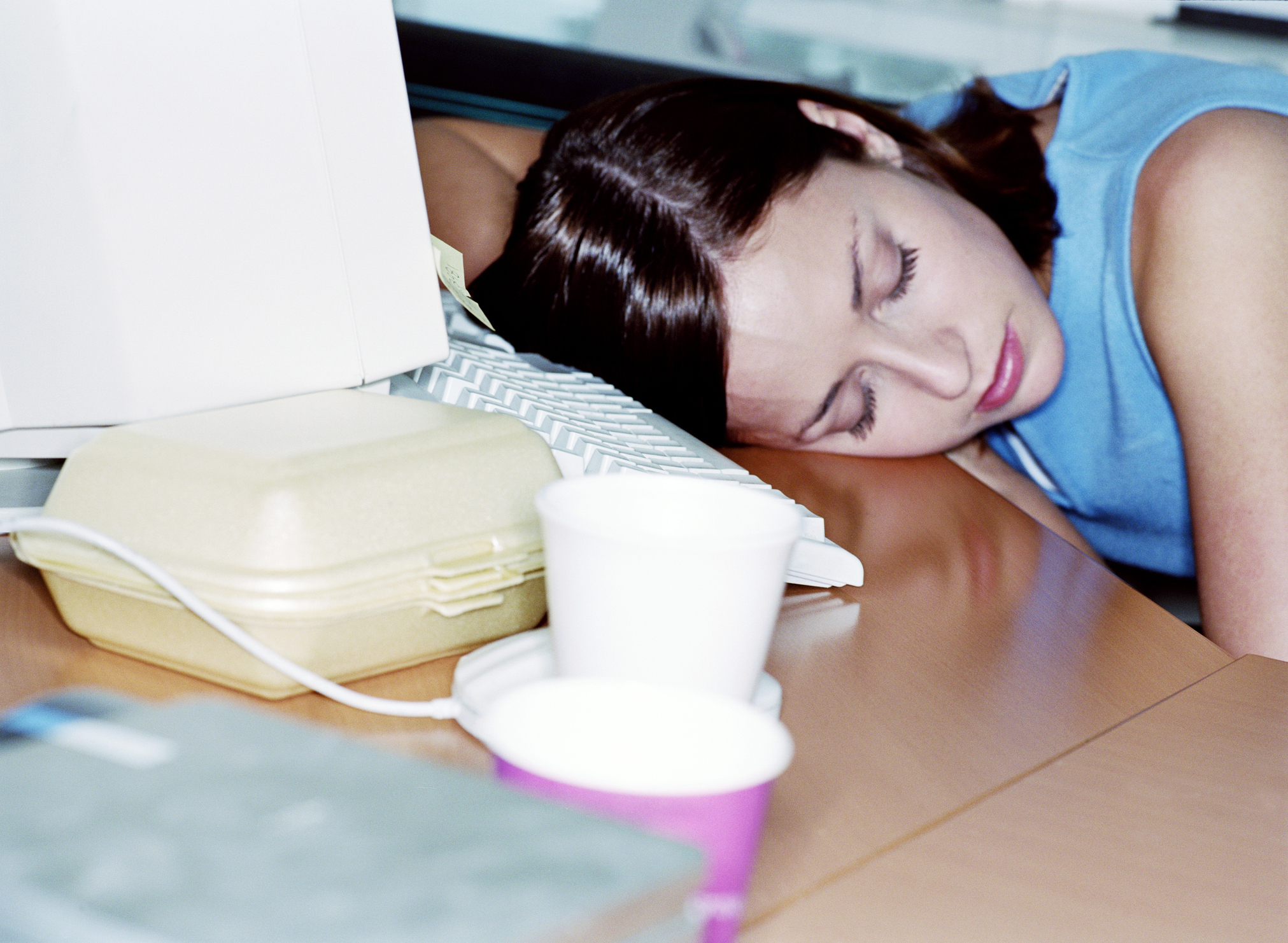 Woman resting head on desk next to computer and foam cups, appearing exhausted or asleep