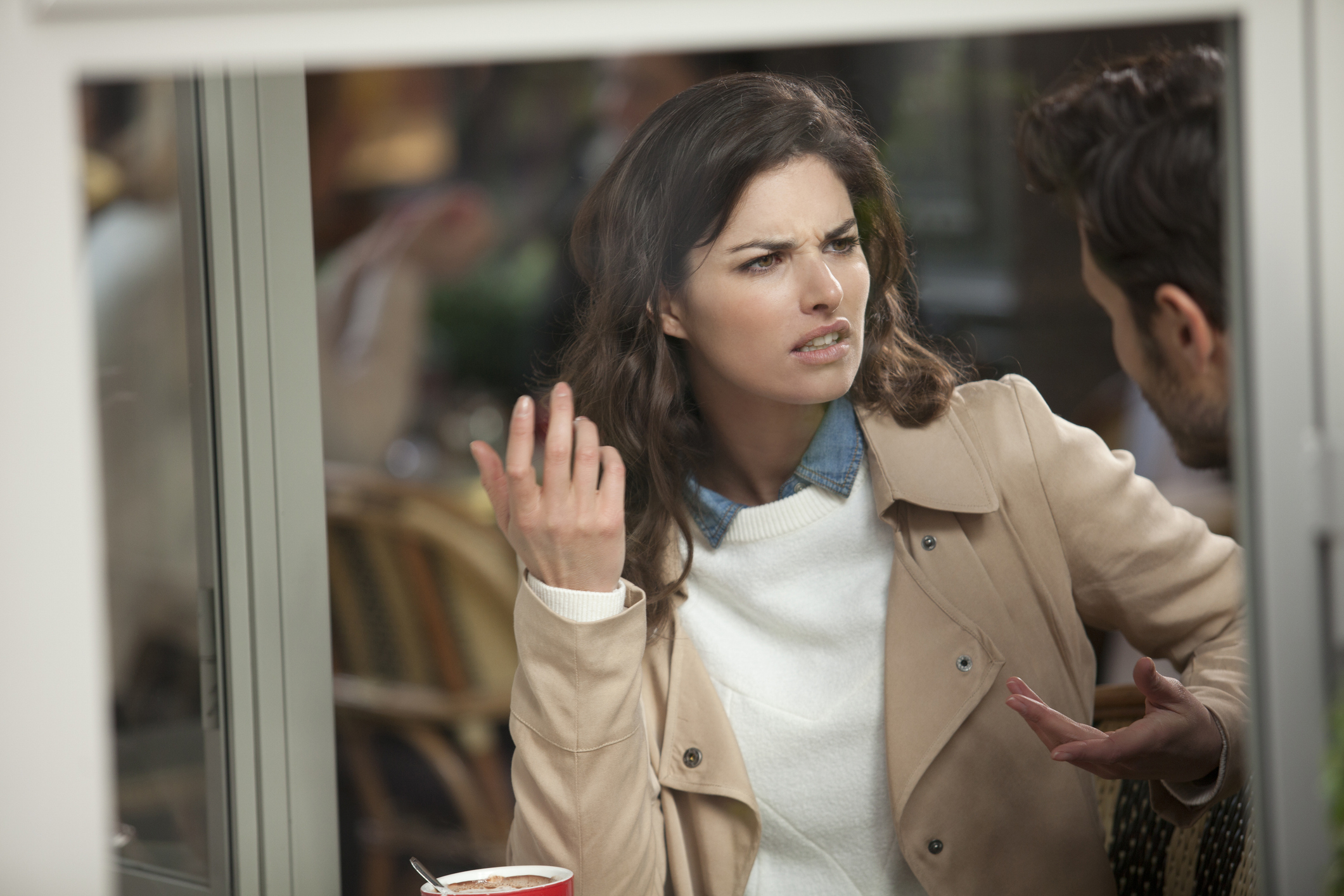 A woman looks annoyed while talking to a man outside a cafe, hand raised in a questioning gesture