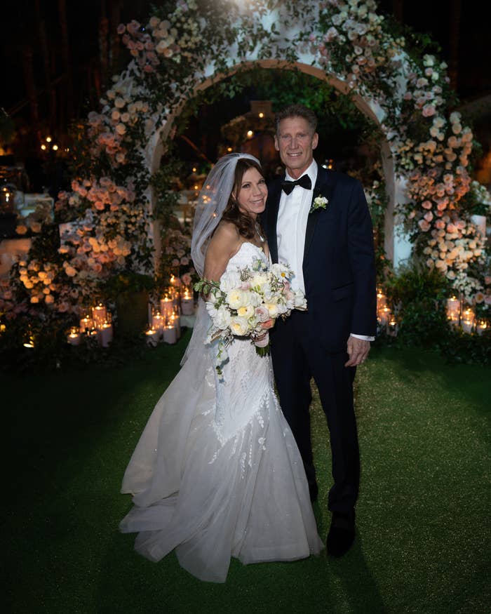 Theresa in a detailed bridal gown and Gerry in a classic suit, stand together in front of their wedding arch decorated with flowers and candles