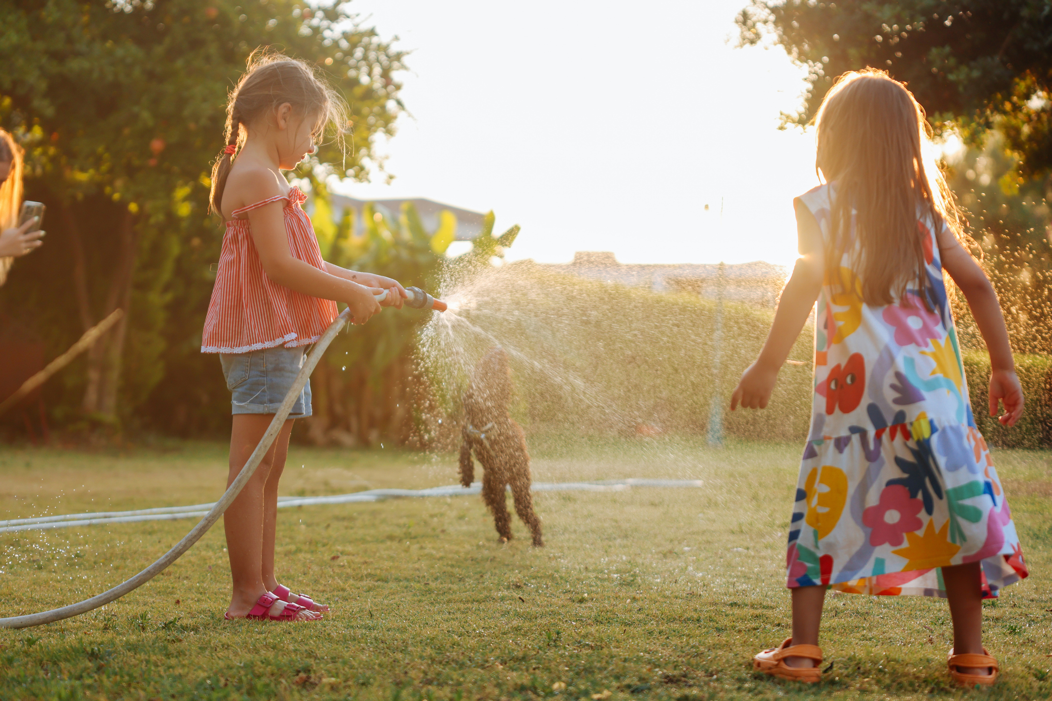Two children play with a hose outdoors; one sprays water while the other watches