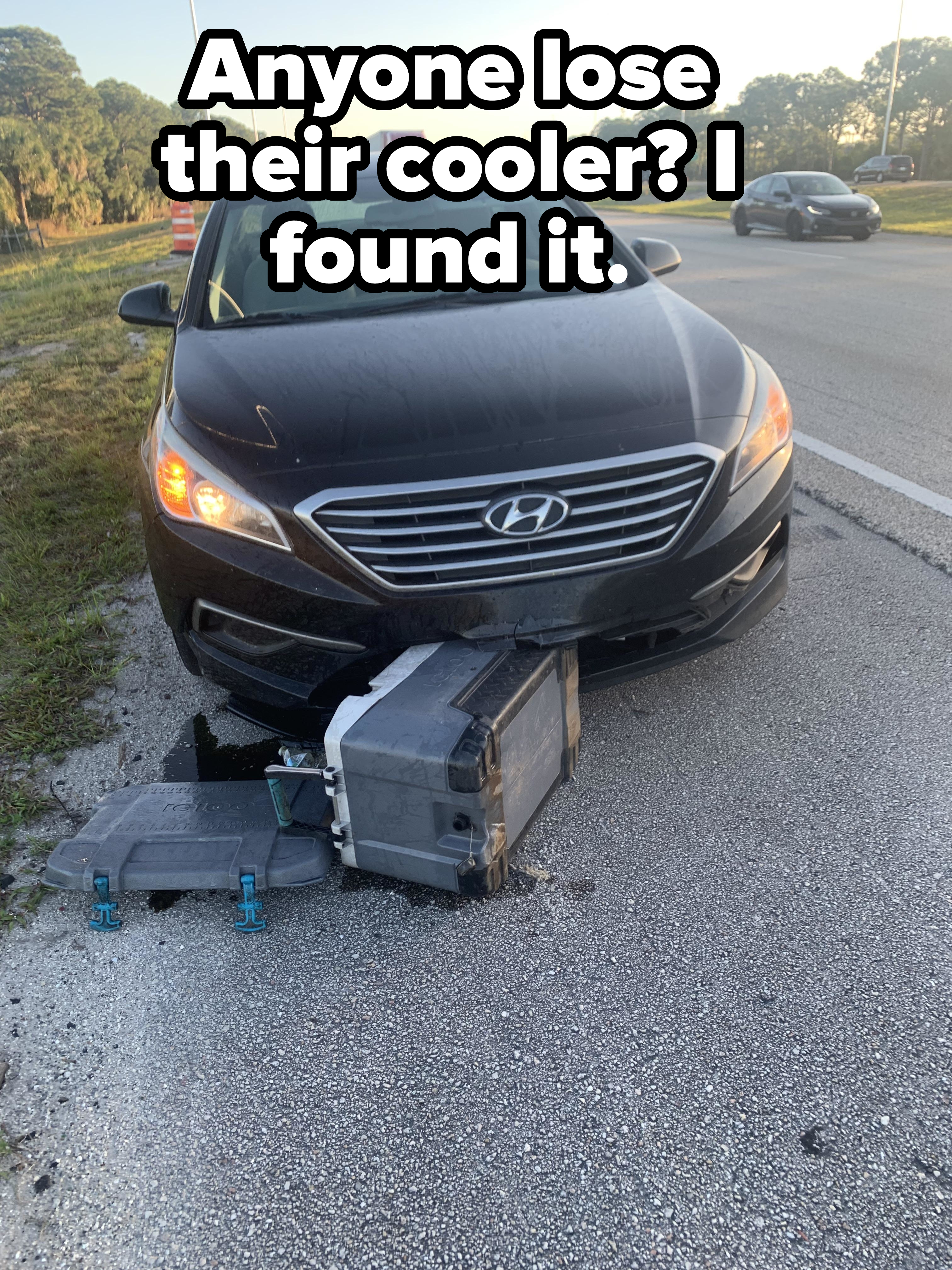 A damaged car with its front tire stuck in a pothole, causing the bumper to partially detach from the vehicle
