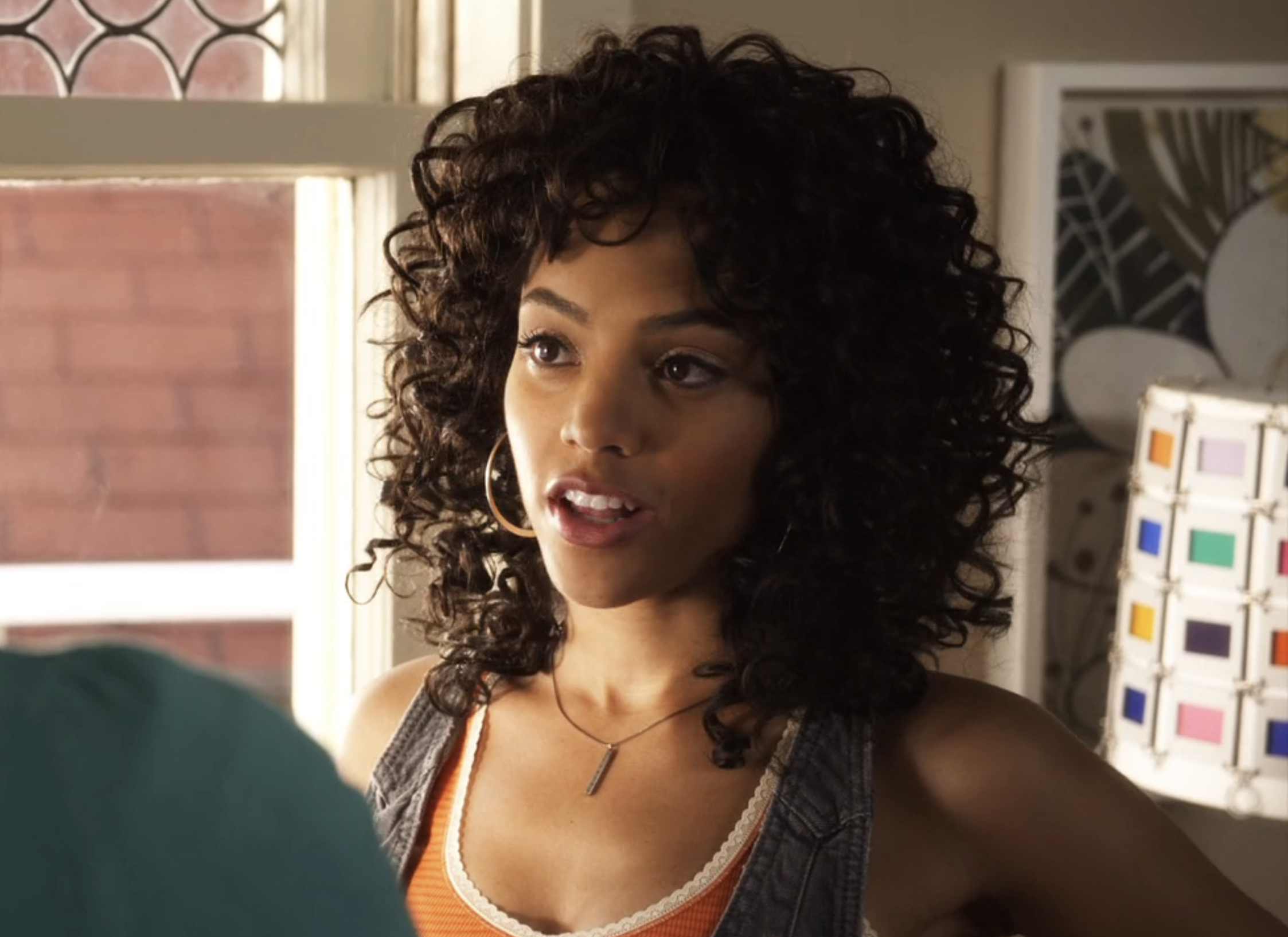 Bianca as Maya with curly hair, tank top, and necklace, indoors near color swatches