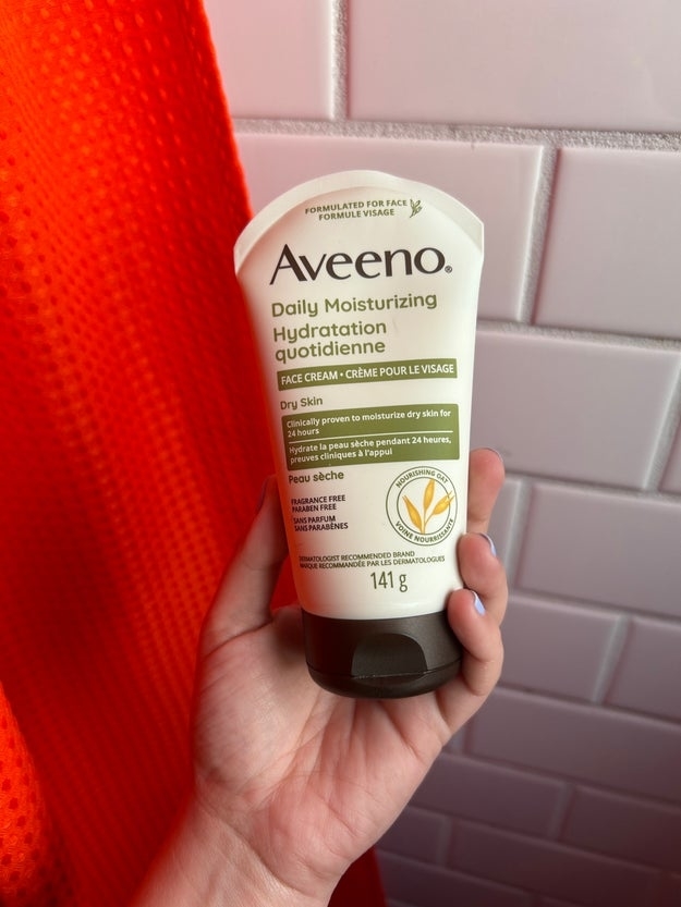 Hand holding Aveeno Daily Moisturizing Face Cream tube against a tiled background