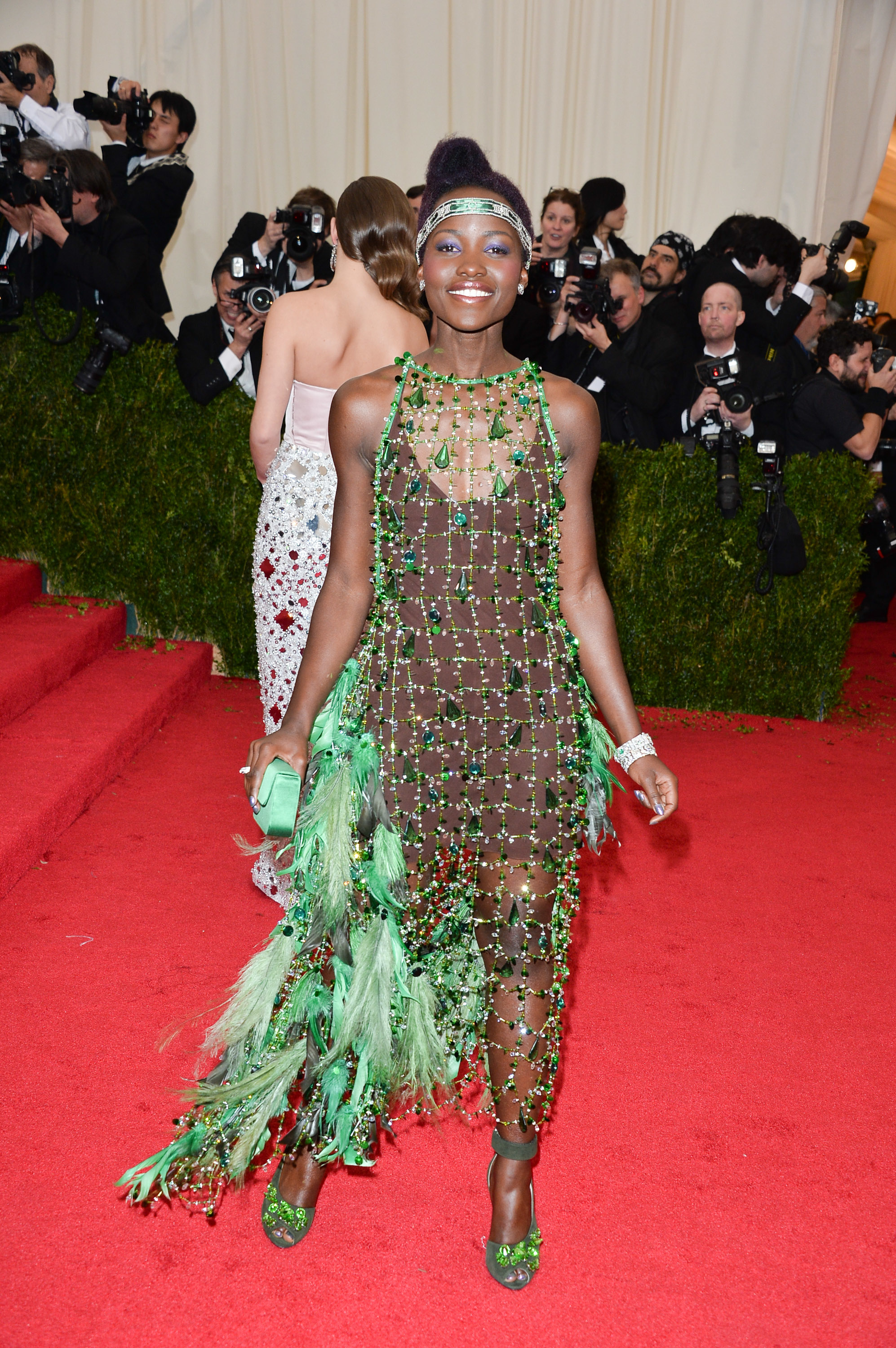 Lupita Nyong&#x27;o in a green beaded dress with feather details, standing on the red carpet with photographers in the background
