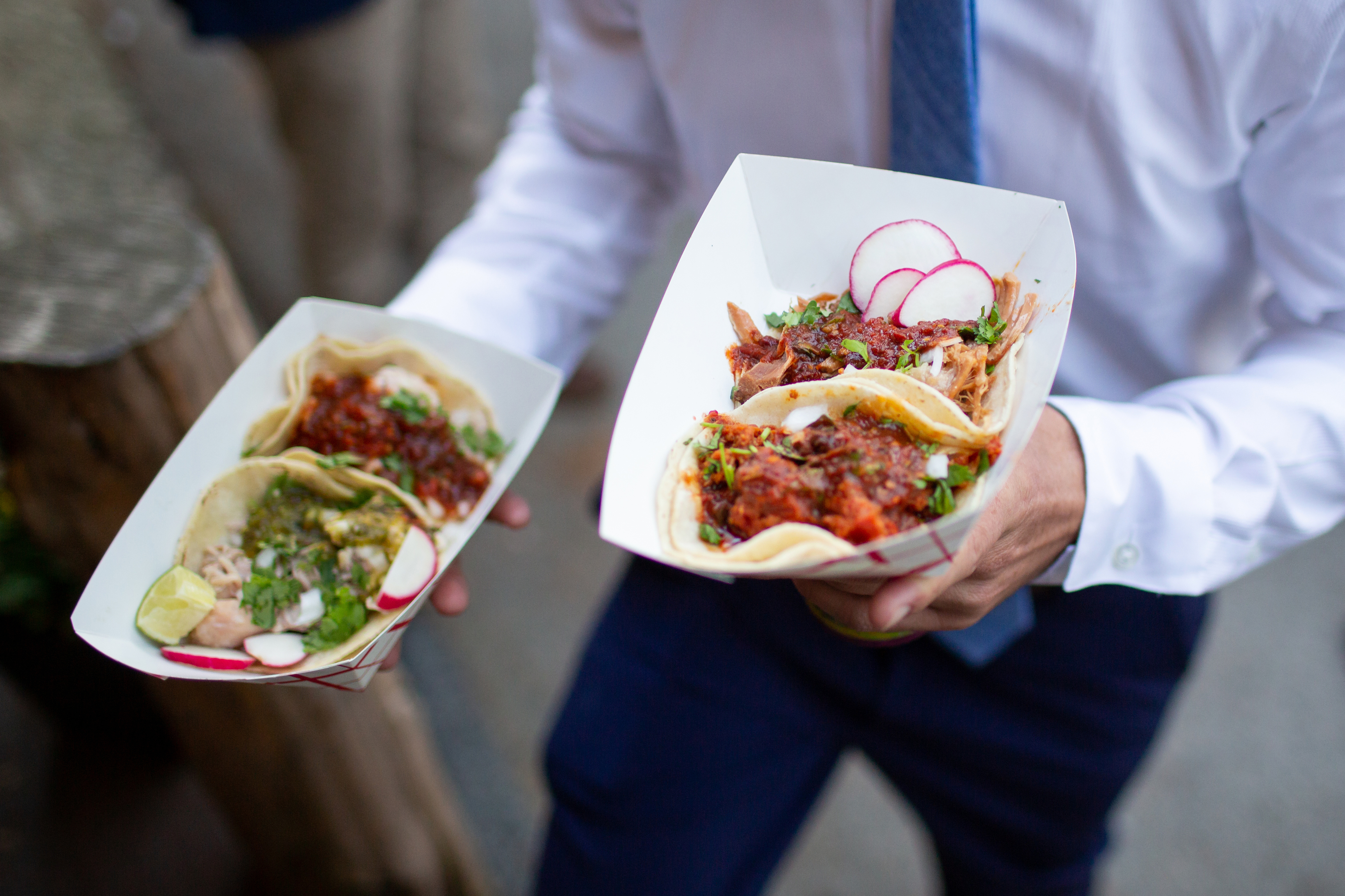 Person holding two tacos with a variety of toppings, likely at a street food stand or outdoor event