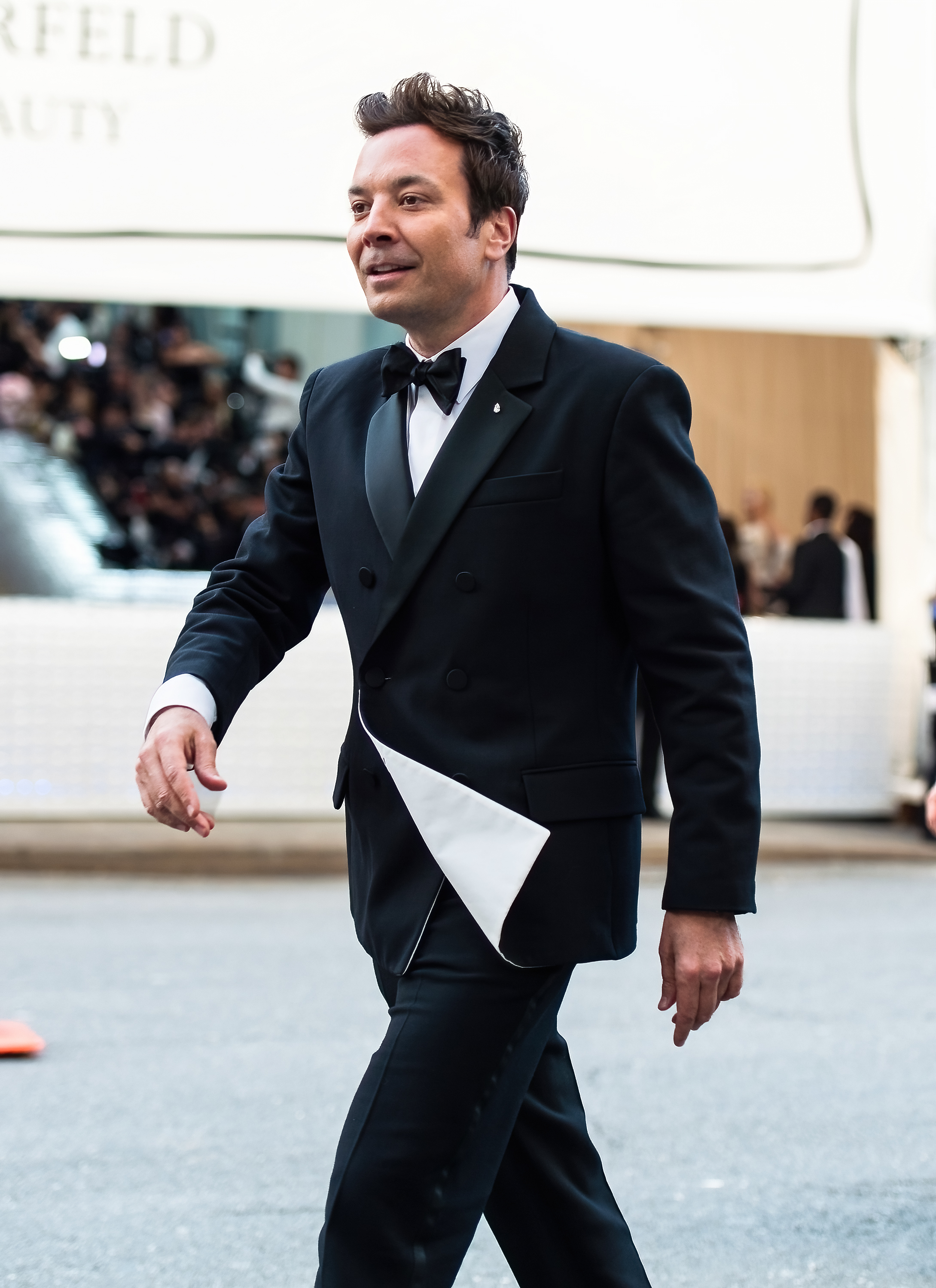 Jimmy Fallon in a black suit and bow tie walking on a sidewalk