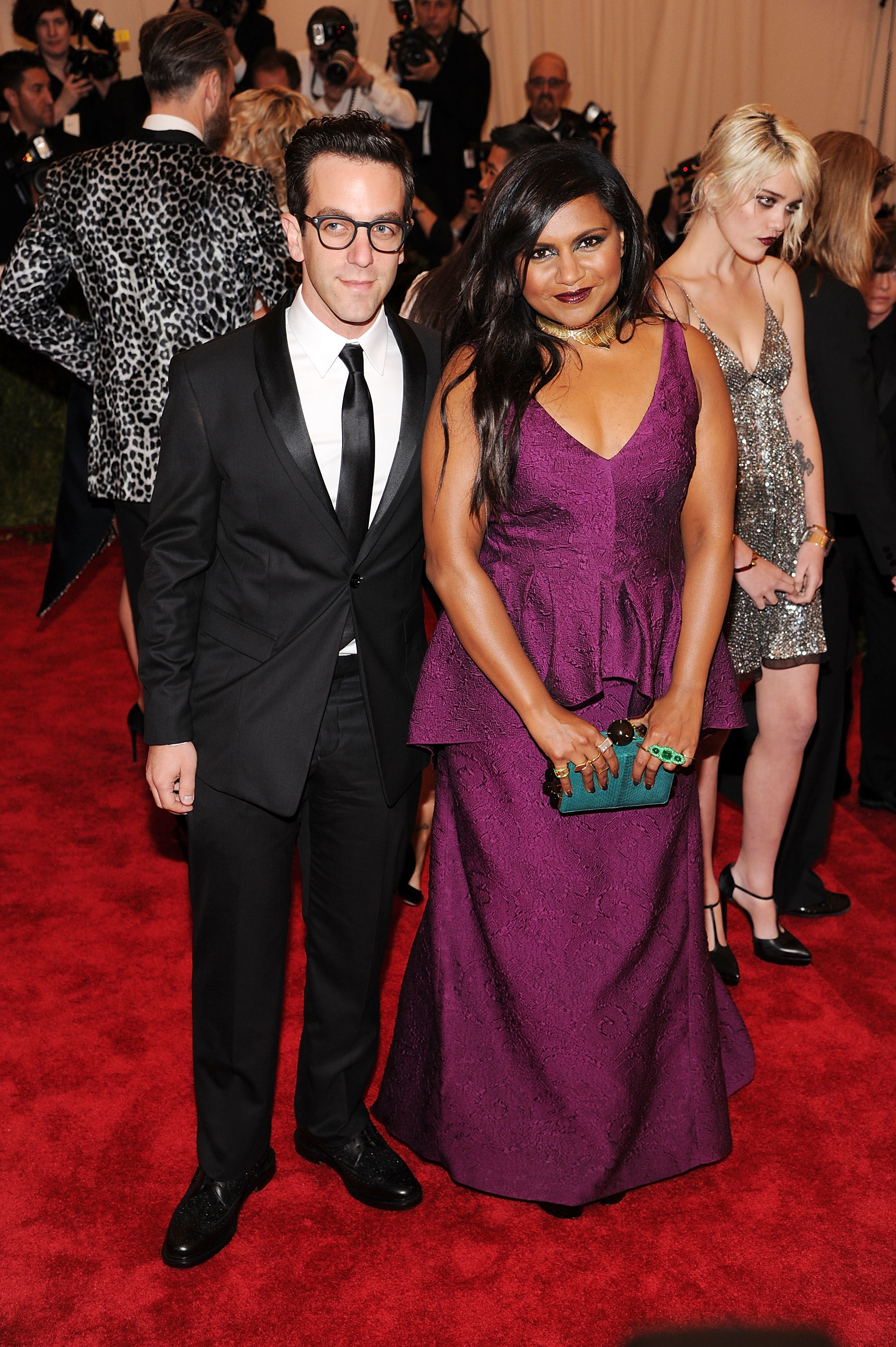 Man in a black suit stands next to a woman in a deep V-neck purple dress with a turquoise clutch at an event