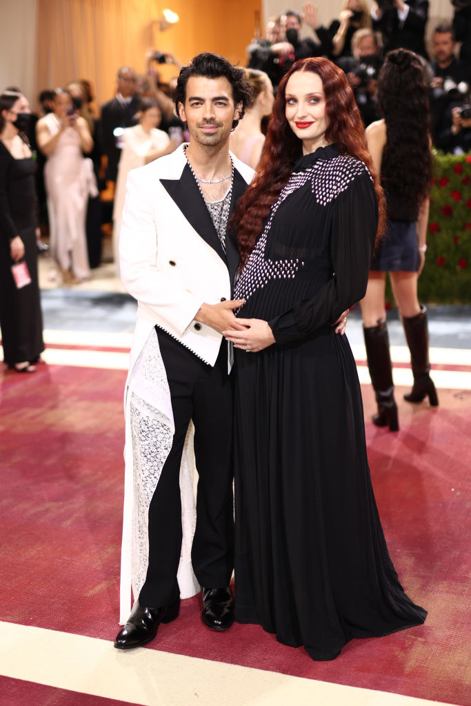 Two celebrities pose together on the red carpet; one in a white and black suit, the other in a black dress with cut-out details