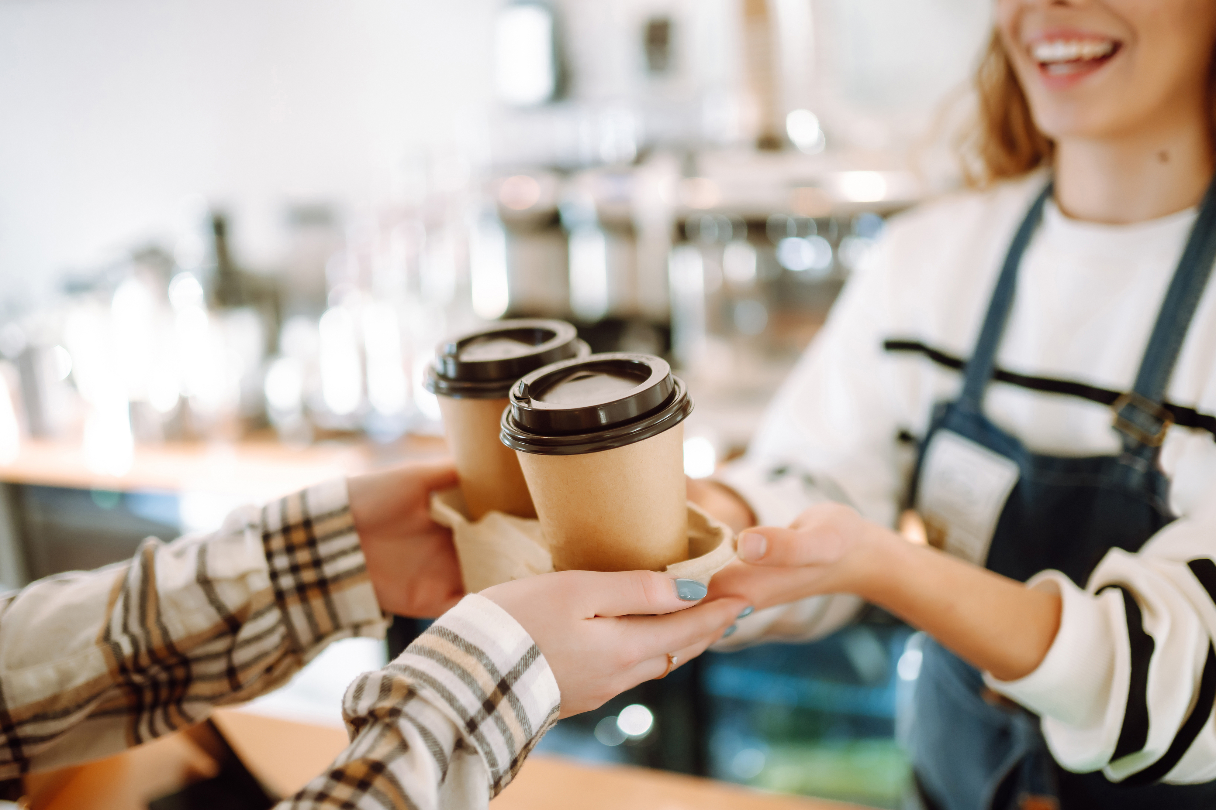 Barista in an apron handing two coffee cups to a customer&#x27;s outstretched hands