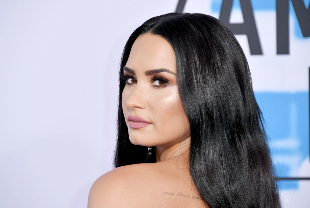 Demi Lovato posing with a side glance, wearing a sleek dress and earrings, tattoo visible on her shoulder