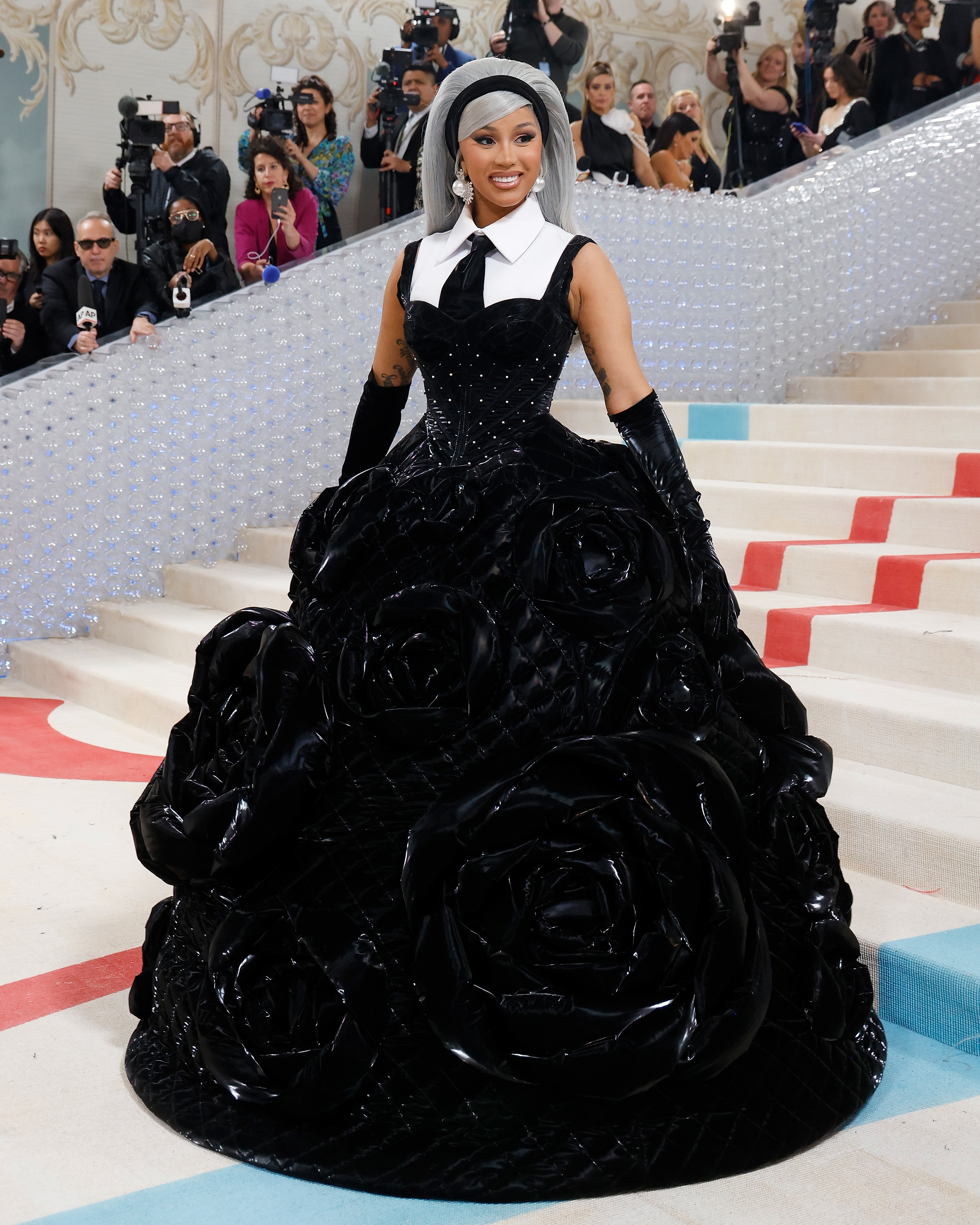 Cardi B in a voluminous black gown with a white collar, posing on stairs at an event