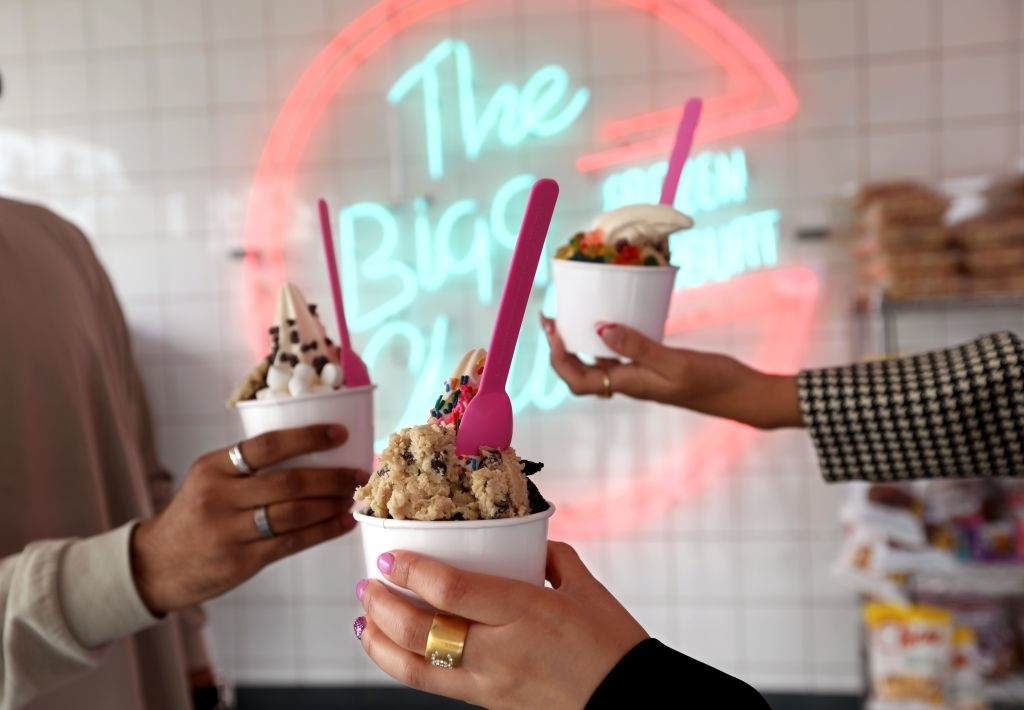 Two people holding cups of ice cream with toppings, neon sign in background reads &quot;The Big O&quot;