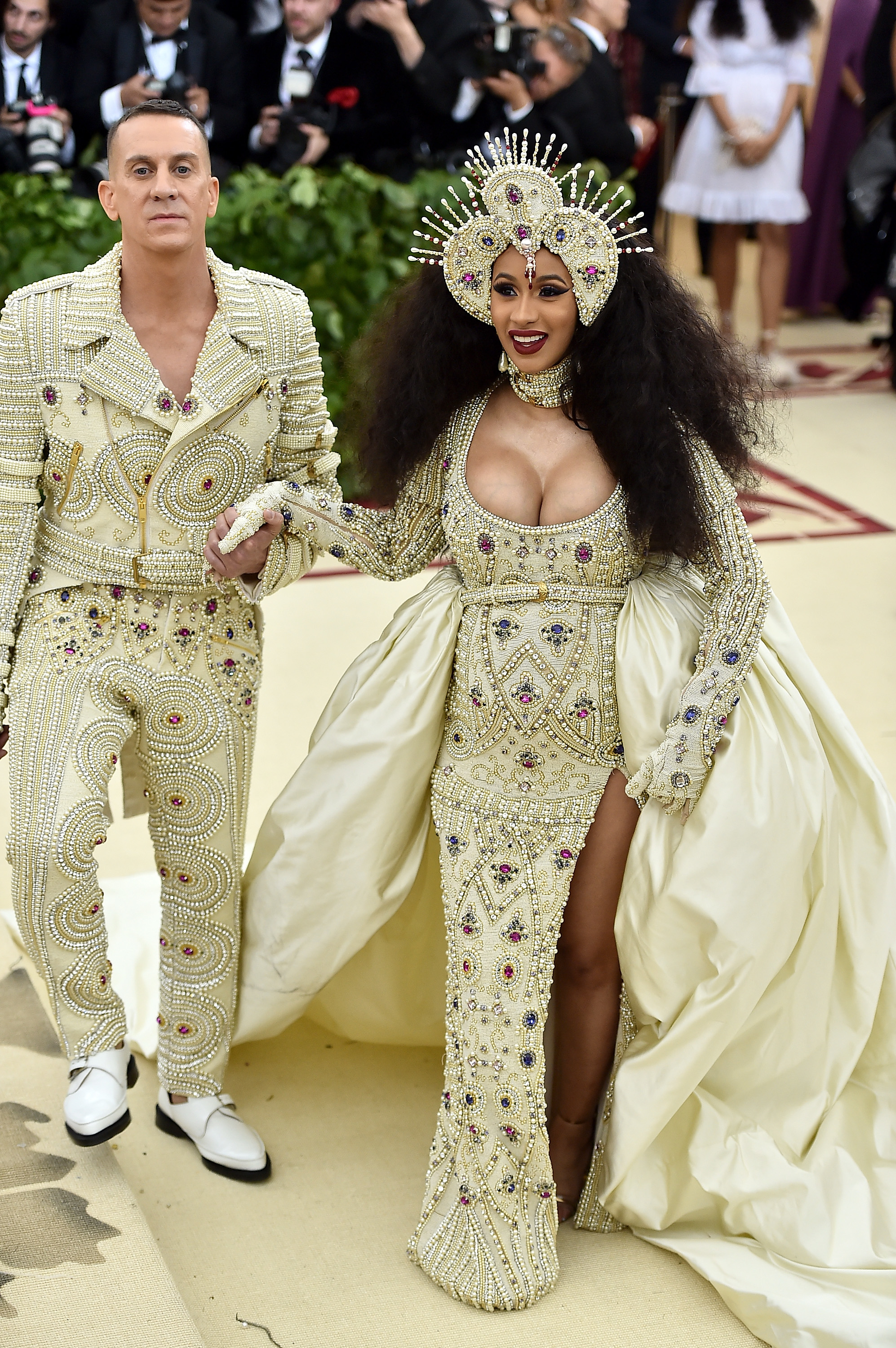 Two people on a red carpet; one in a bejeweled suit, the other in an ornate headdress and gown with a high slit