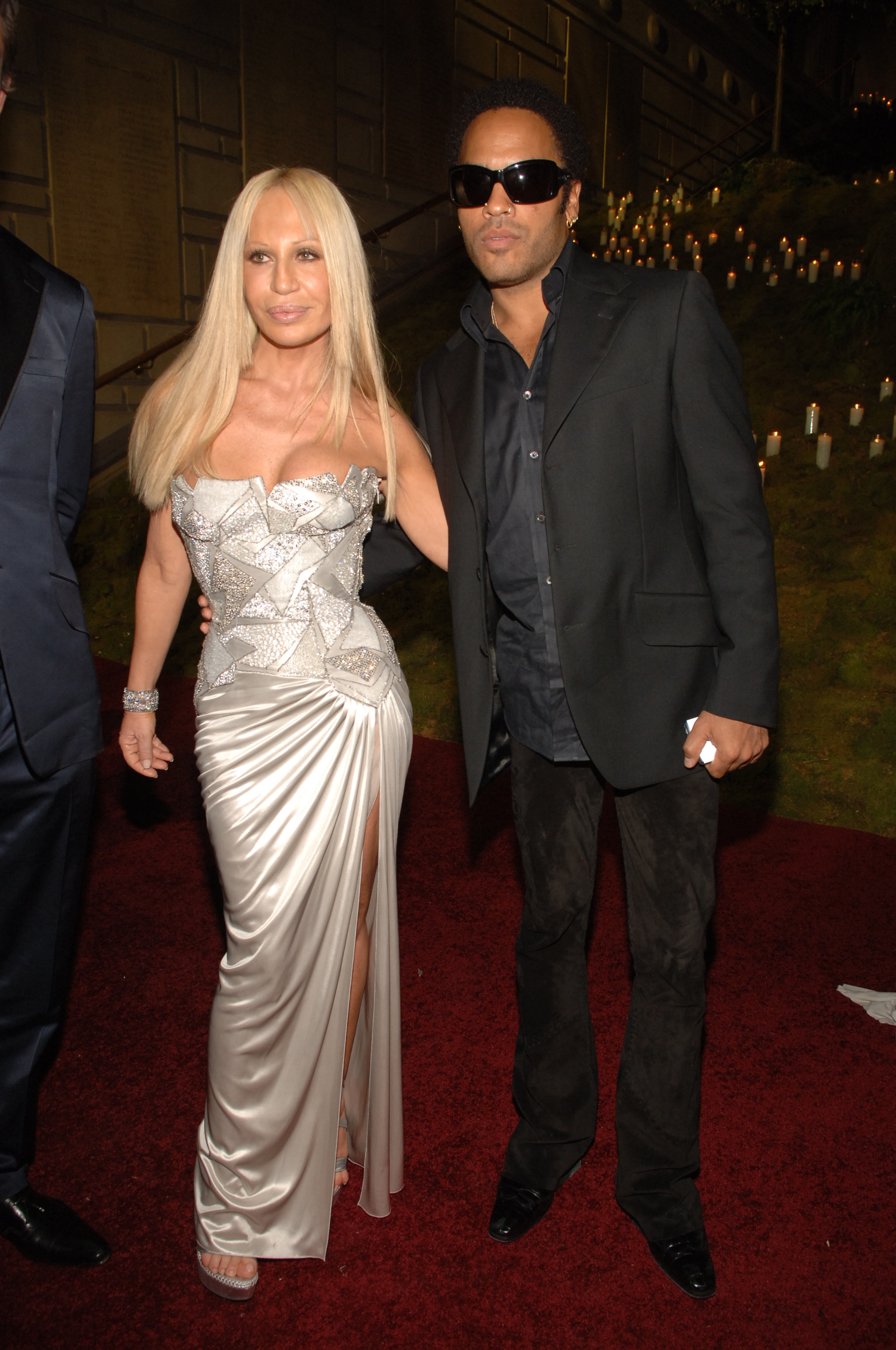 Two celebrities on red carpet, woman in embellished silver gown with slit, man in black suit and sunglasses