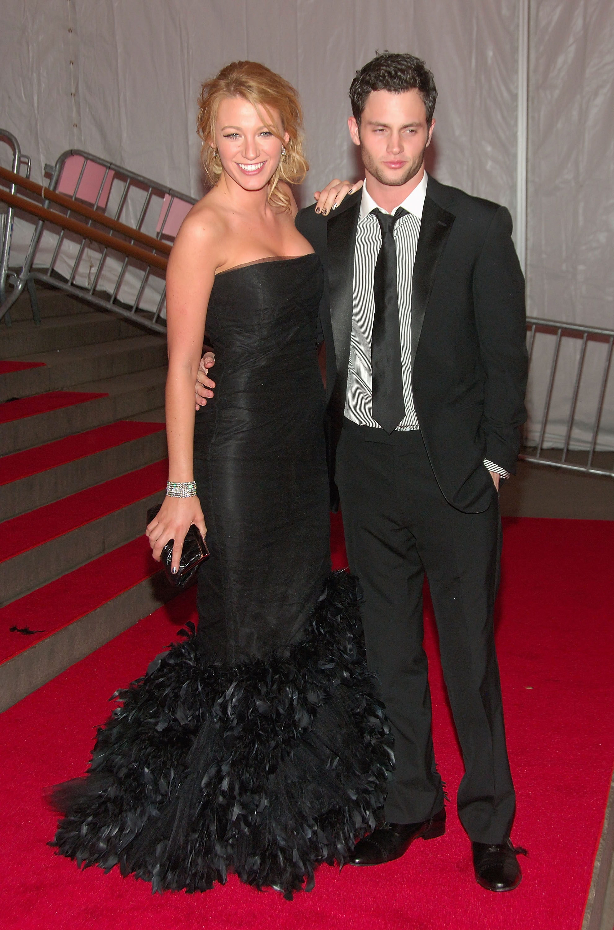 Two celebrities on red carpet; one in a black strapless gown with feather details, the other in a classic black suit