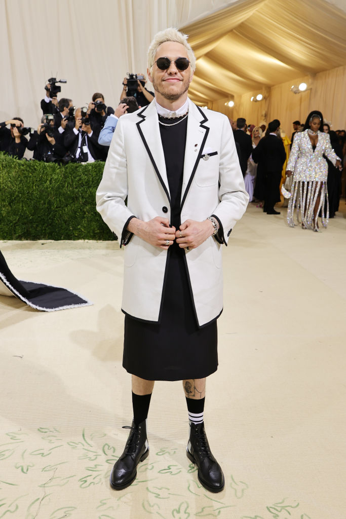 Person in a tuxedo jacket, shorts, and boots, standing on a carpet, with photographers in the background