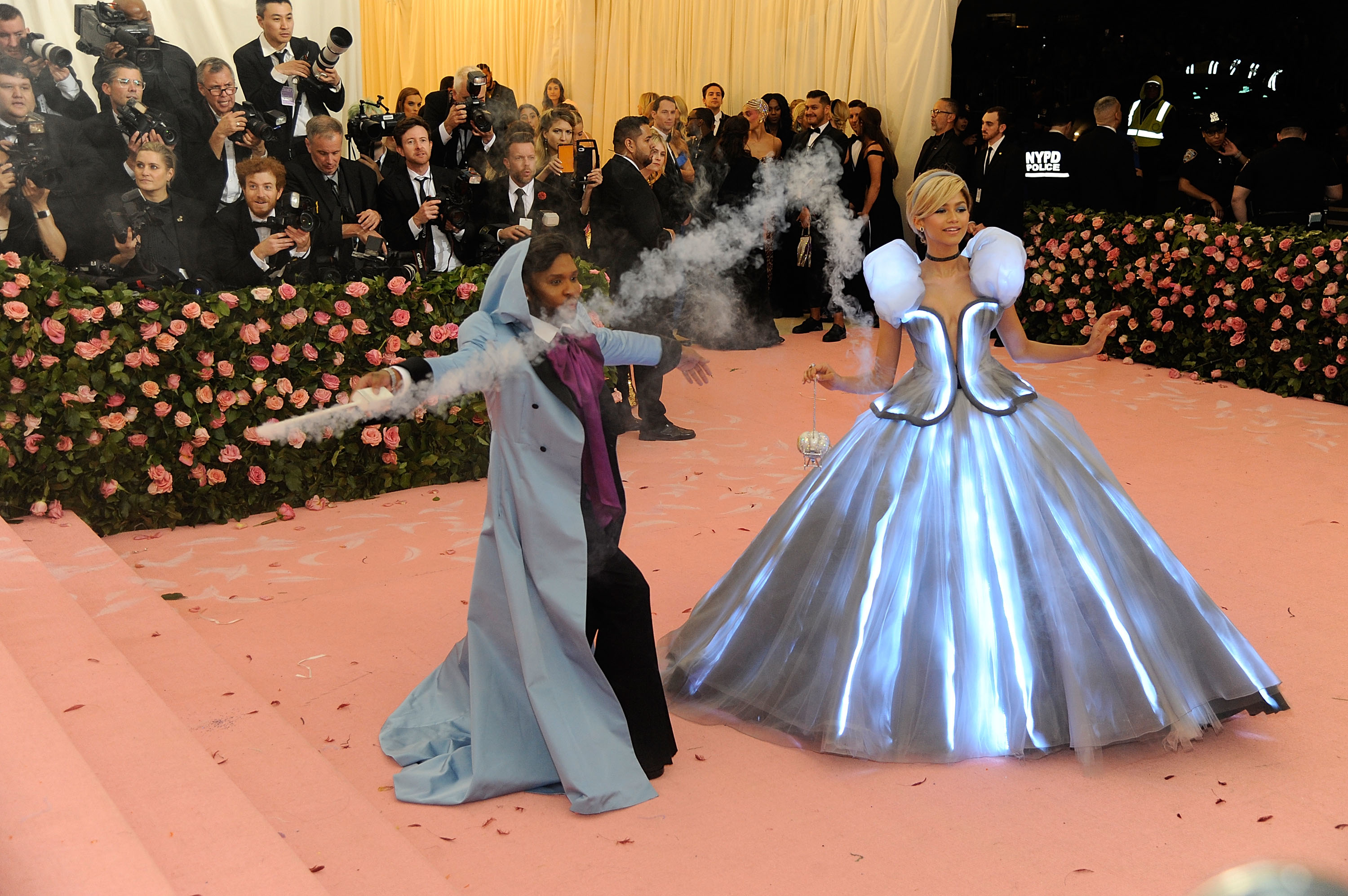 Person in a futuristic silver gown with a smoke effect alongside a person in blue, both on a carpet event