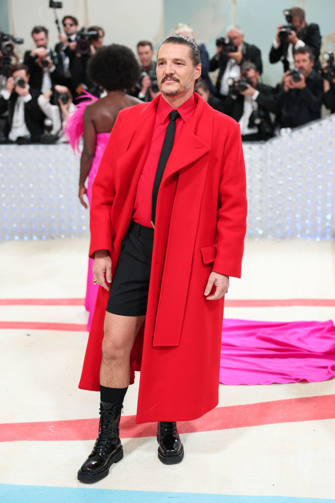 Man in a red coat and black shorts standing on event carpet, photographers in background