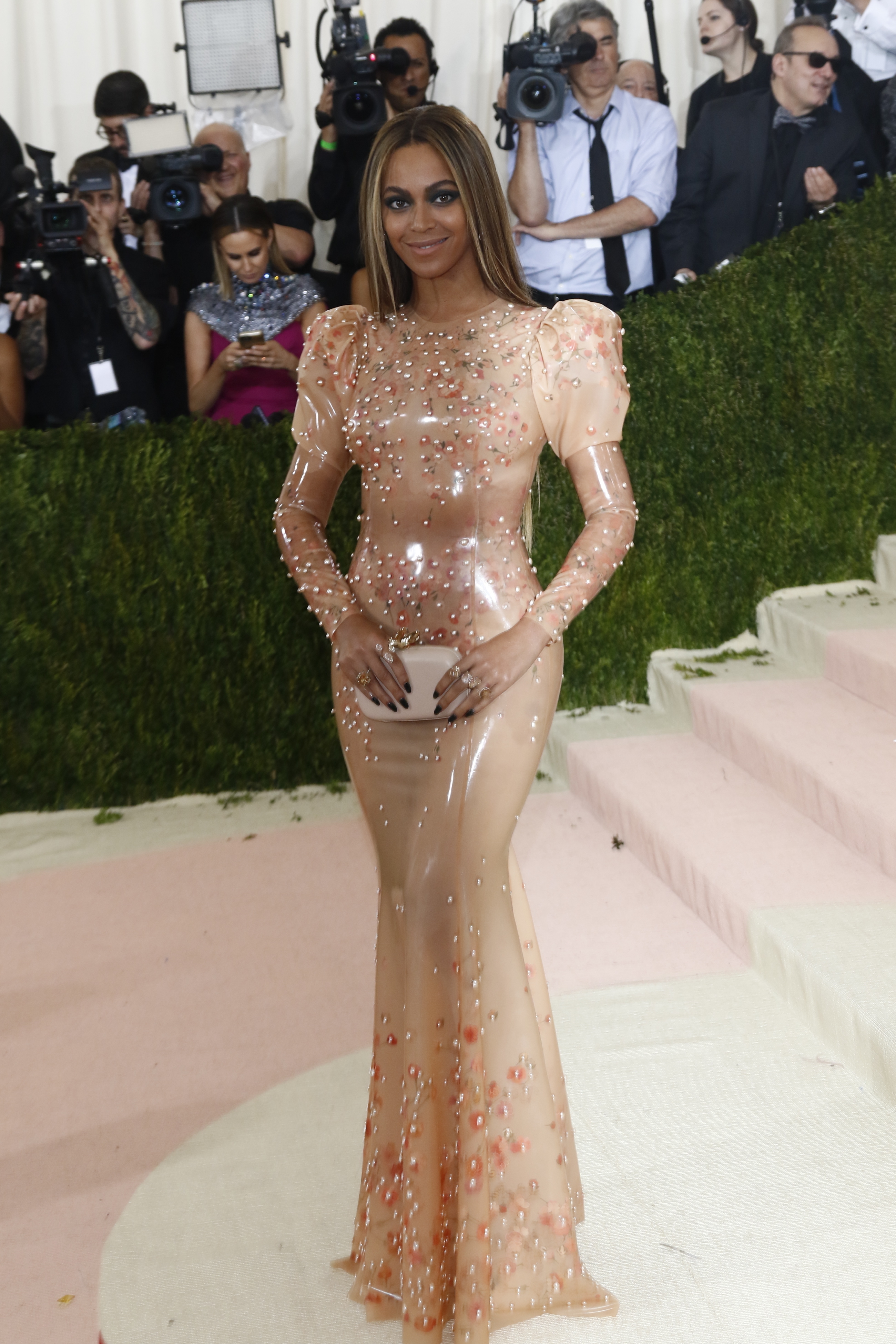 Beyoncé in a sheer, embellished gown at an event, with photographers in the background