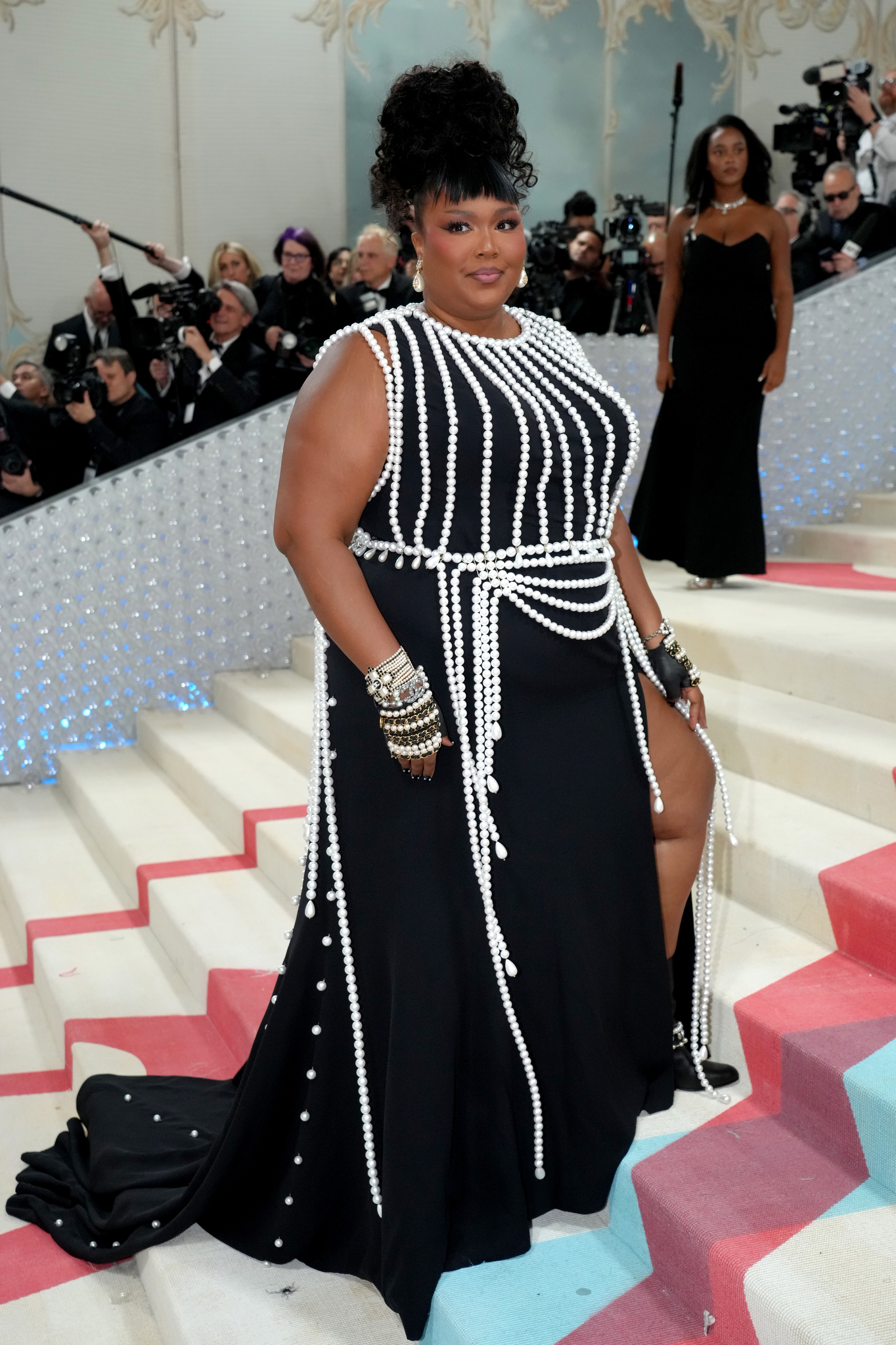 Lizzo stands on stairs in a black dress with white beaded embellishments, bracelets, and updo hairstyle