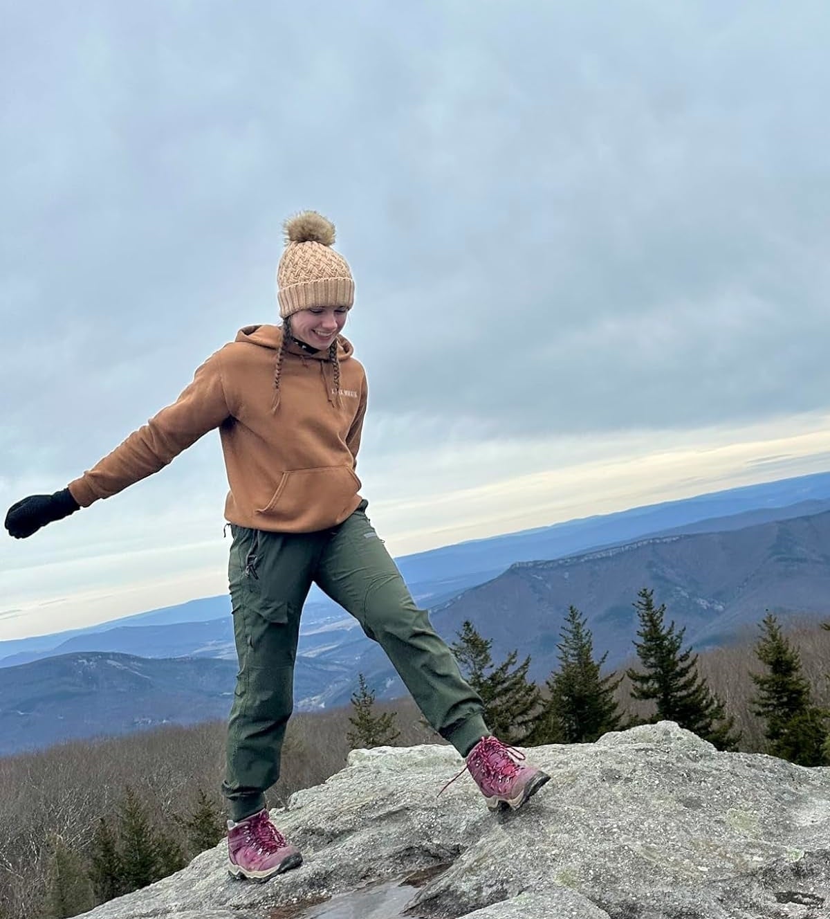 A reviewer in hiking pants with a beanie balancing on a mountain rock with a scenic view in the background