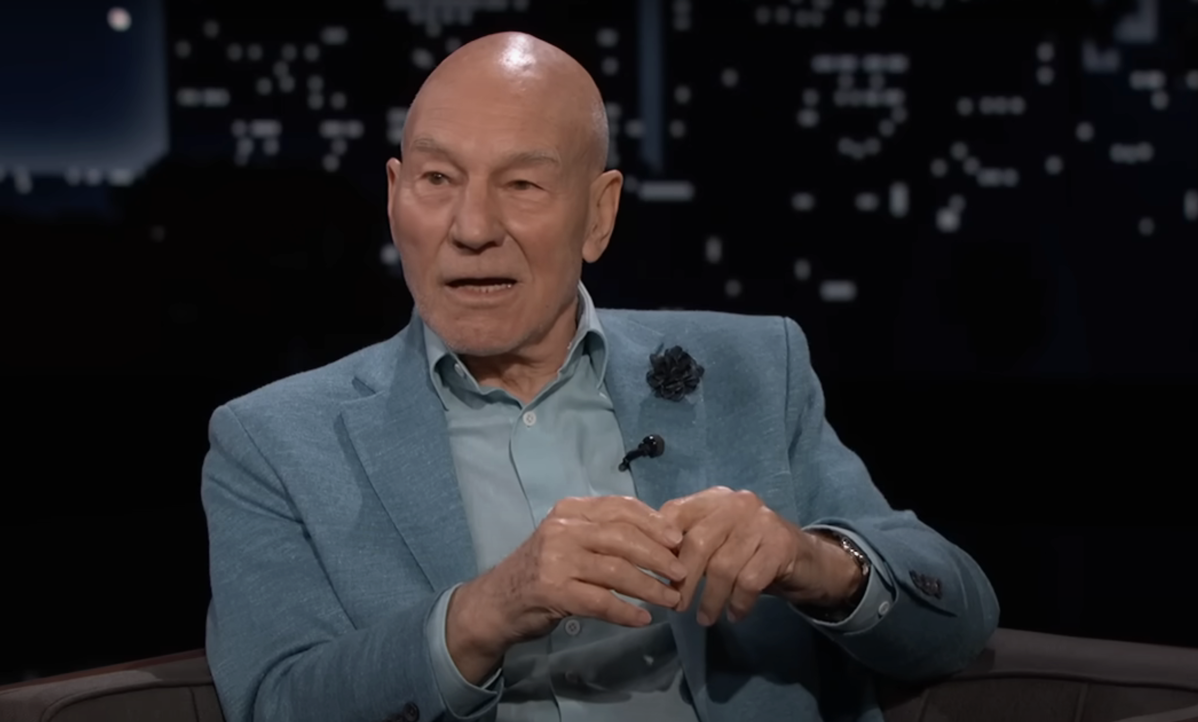 Patrick Stewart sitting, wearing a light blue suit, on a talk show with a cityscape backdrop