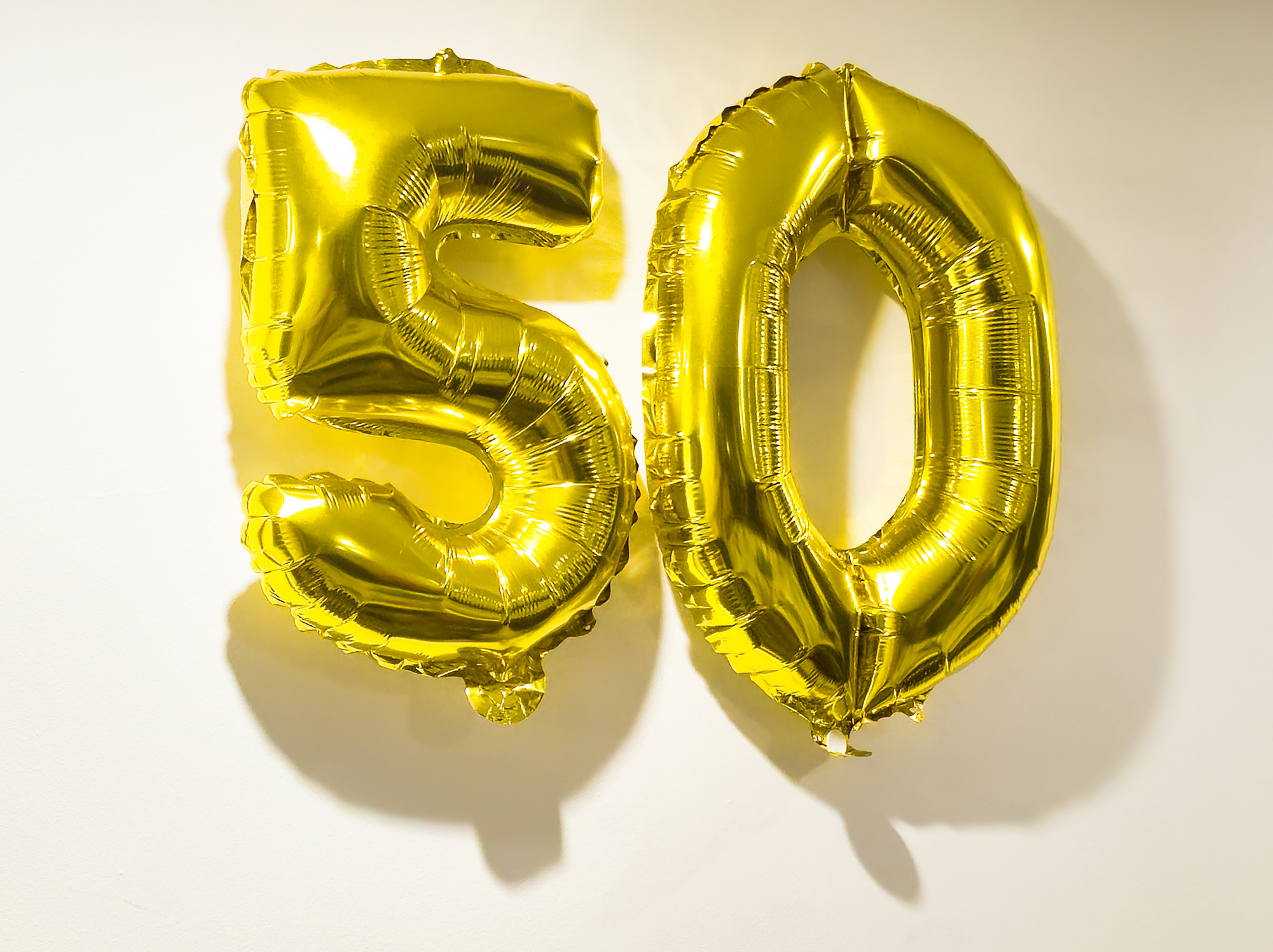 Two gold foil balloons shaped as the number 50 against a plain wall, likely symbolizing a milestone
