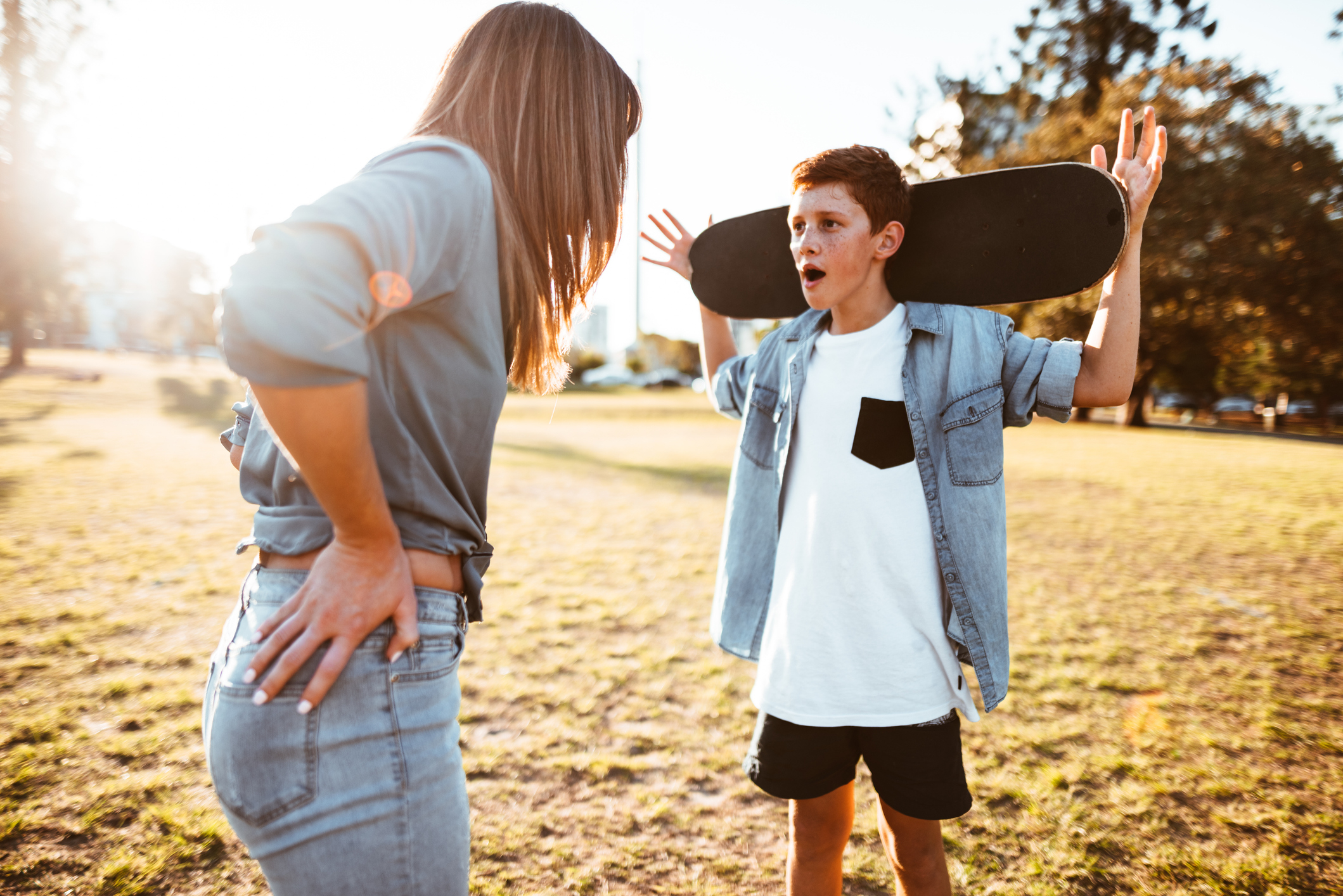 A mother speaking to her son who is holding a skateboard in a park