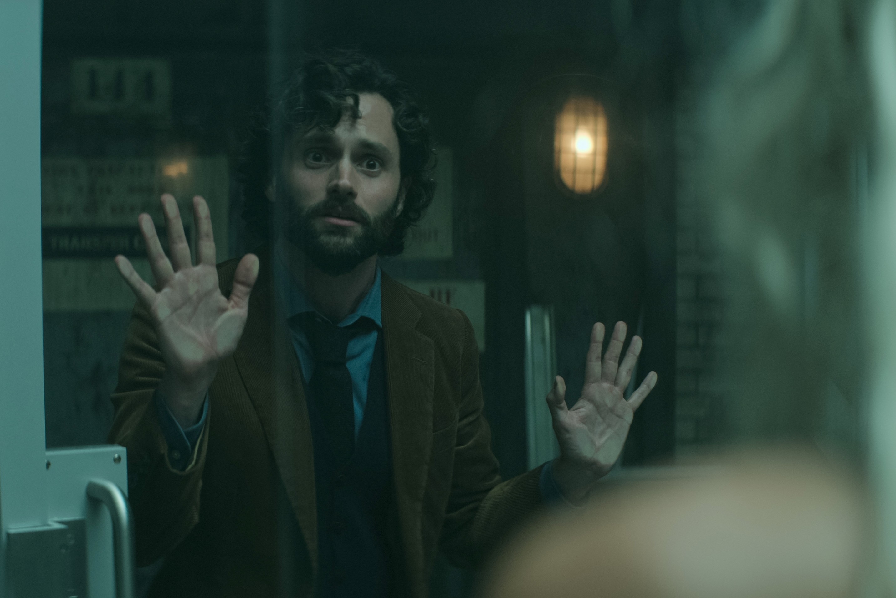 Penn Badgley in a suit with hands up against a window, showing a surprised expression
