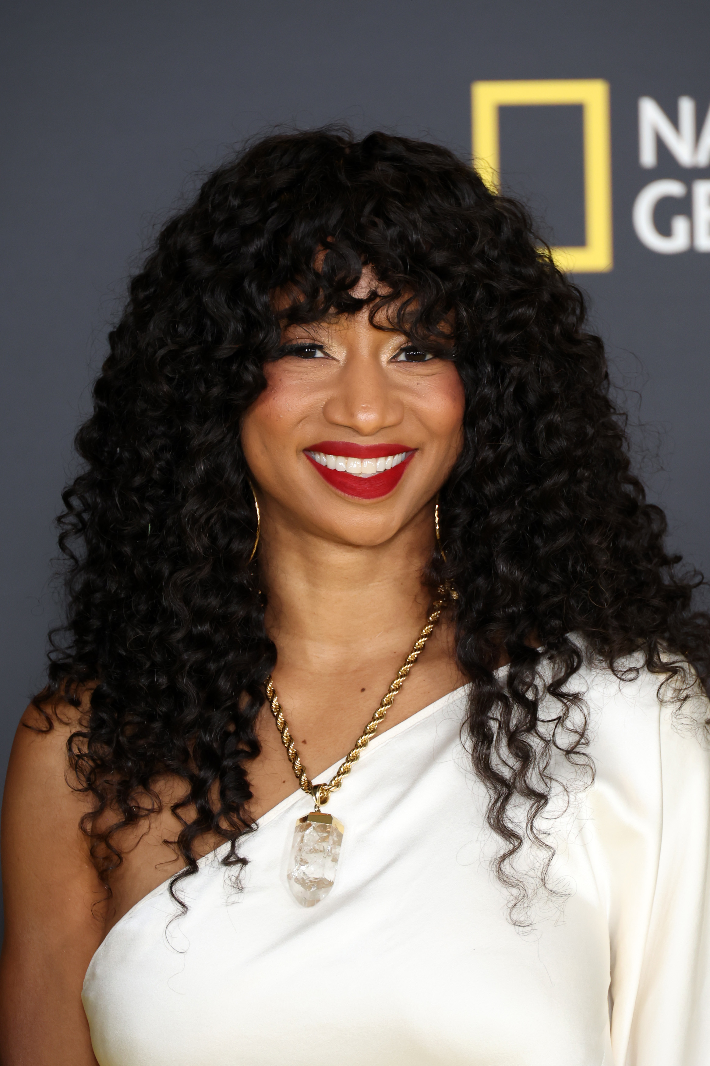 Smiling person on red carpet in white top with gold necklace, curly hair, and red lipstick