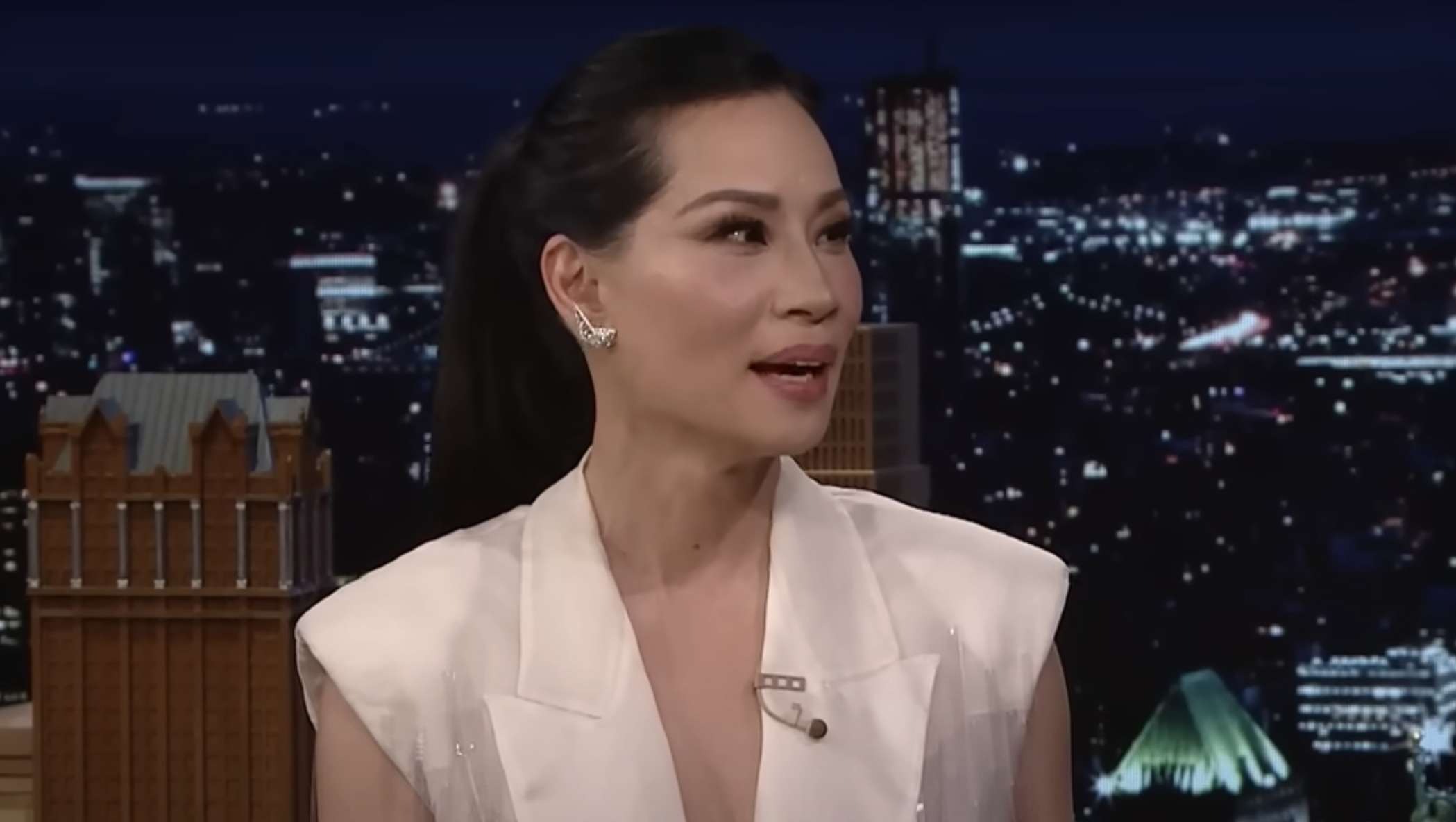 Woman on talk show wearing a v-neck layered lapel blouse, earrings, with cityscape background