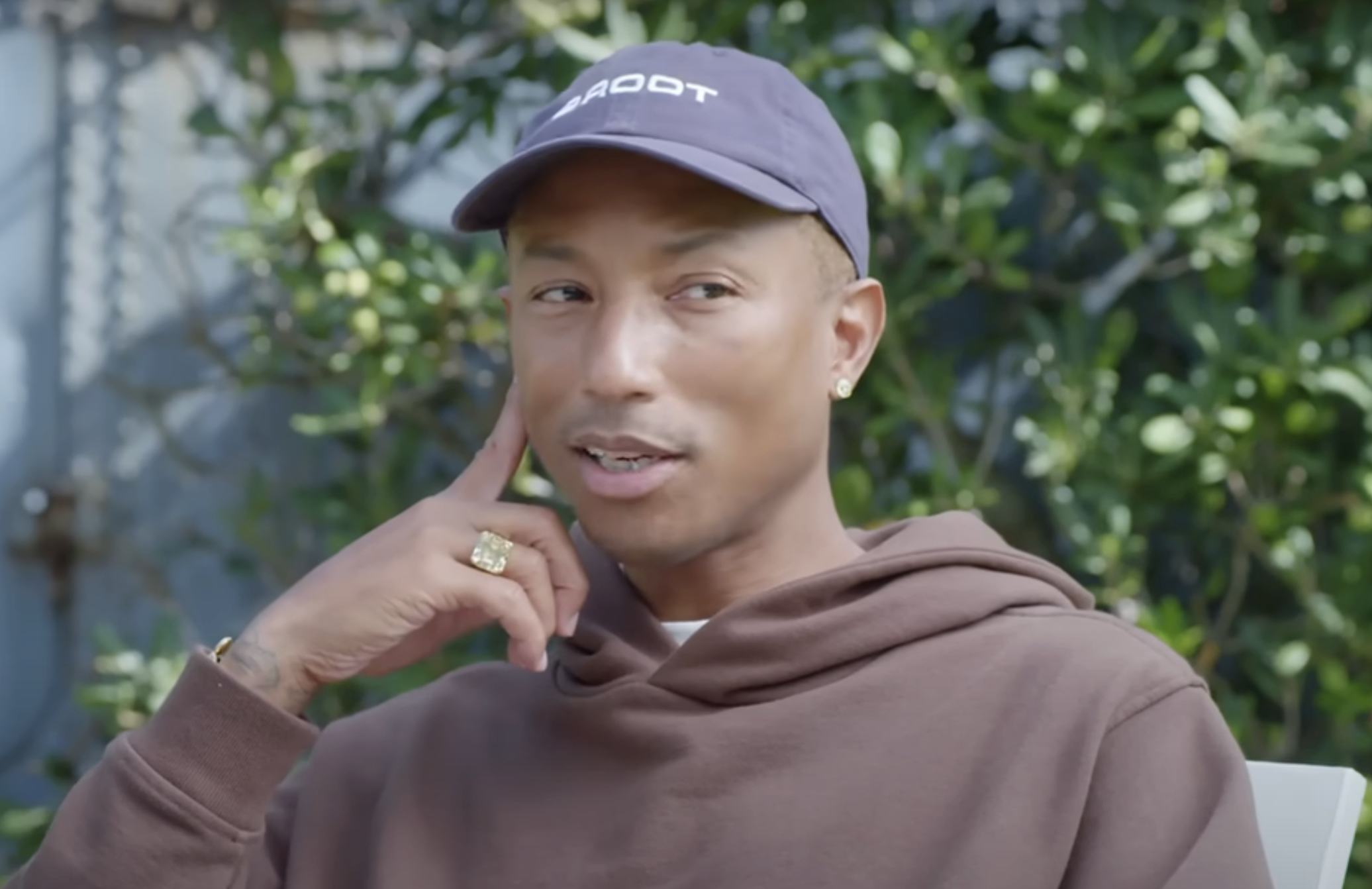 Pharrell Williams in a casual hoodie and cap, seated outdoors, smiling slightly with hand on chin