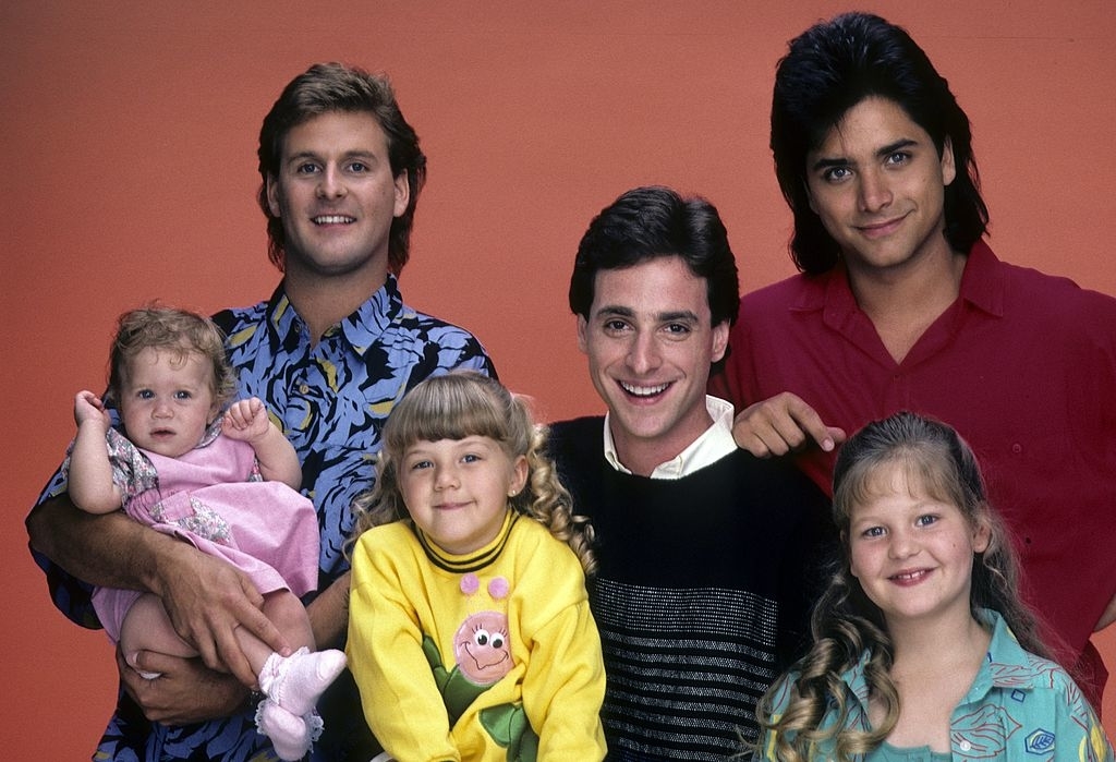 Cast of &quot;Full House&quot; with three men standing and three young girls, two sitting and one held, in casual clothes