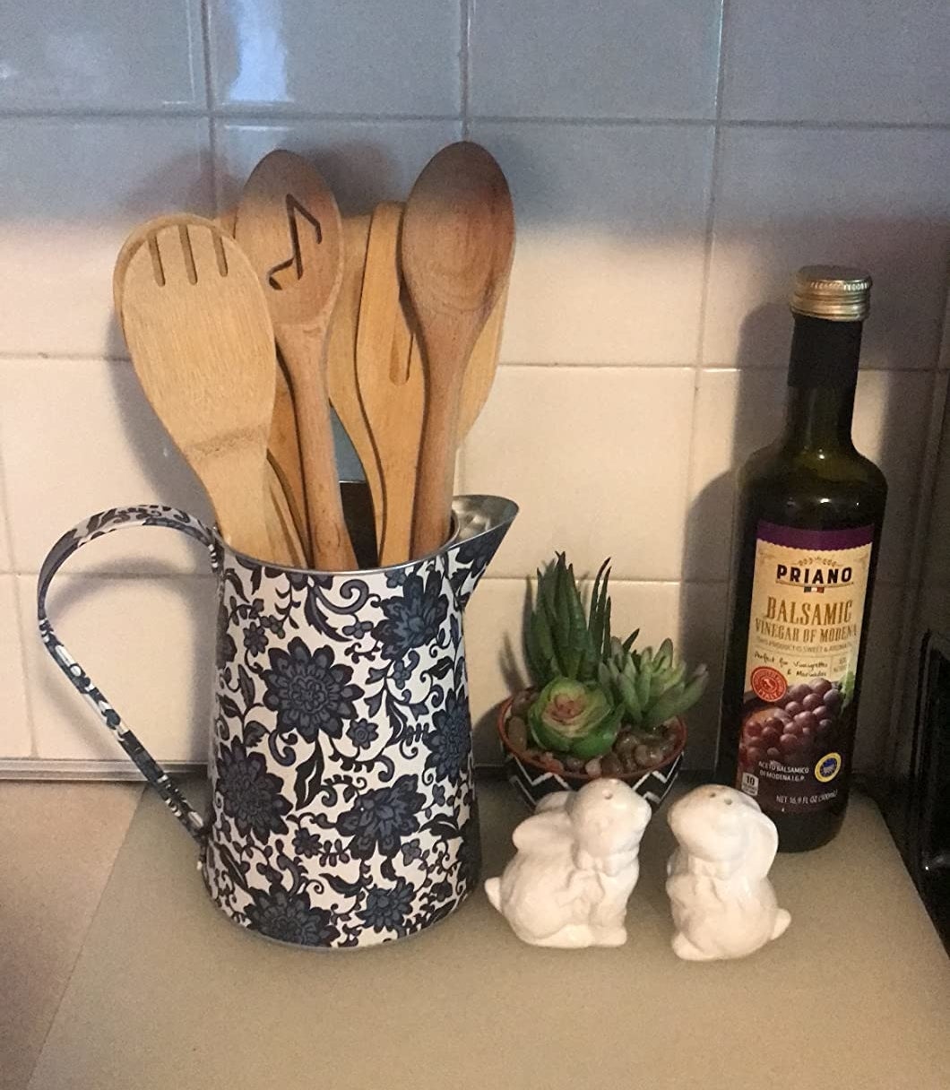 A pitcher with floral patterns holding wooden utensils next to a bottle of balsamic vinegar and decorative items on a kitchen counter