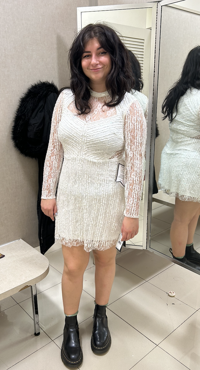 Person in a fitting room wearing a white lace dress with sheer sleeves and black boots, standing in front of a mirror