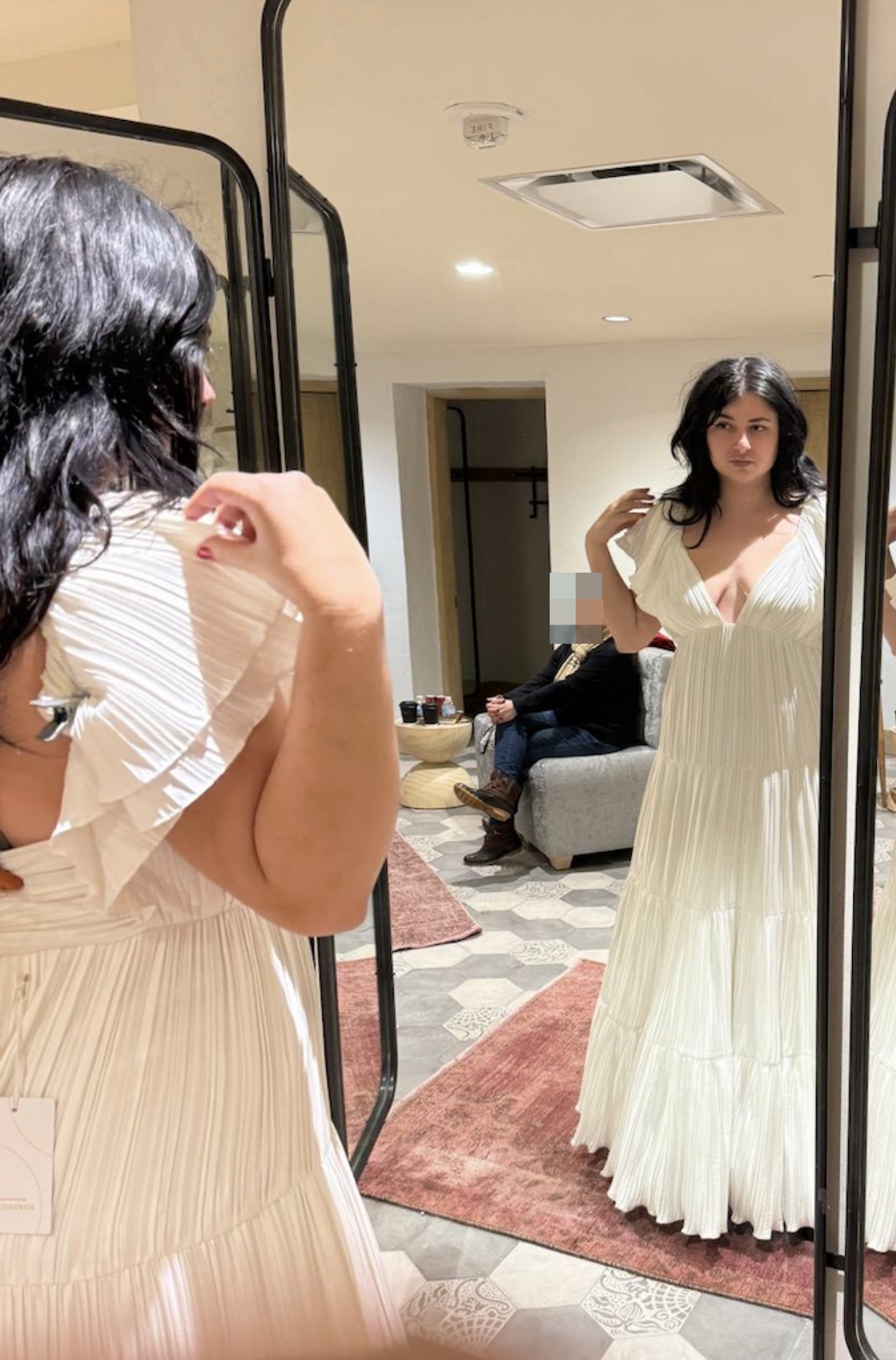 A person tries on a pleated dress in a mirror, another person seated in background