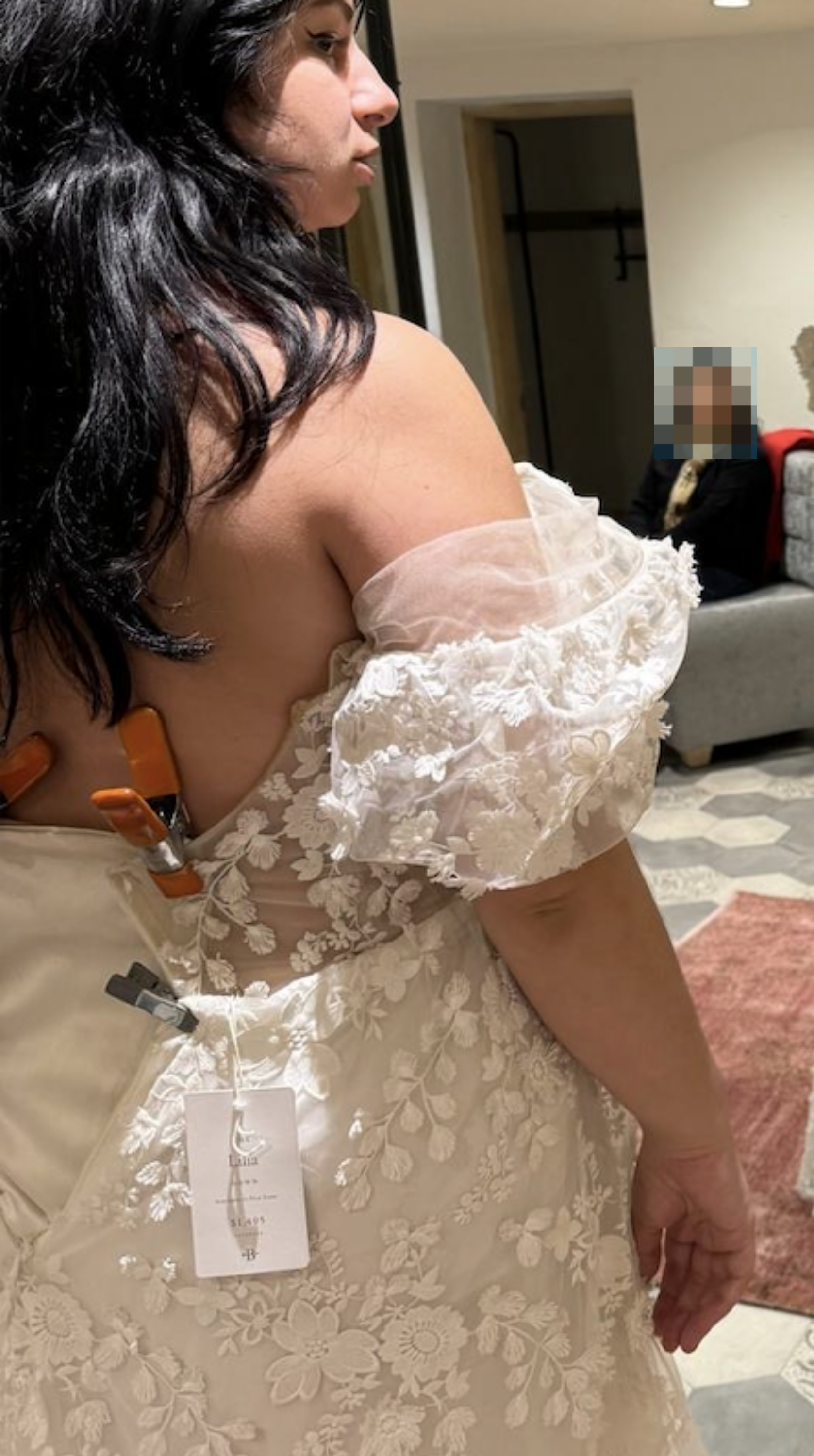 Person in a bridal shop trying on a lace dress with off-shoulder sleeves, another person seated in background