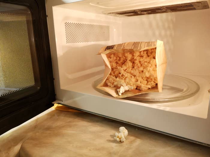 An opened bag of popcorn has overflown in a microwave, with some kernels spilled onto the turntable