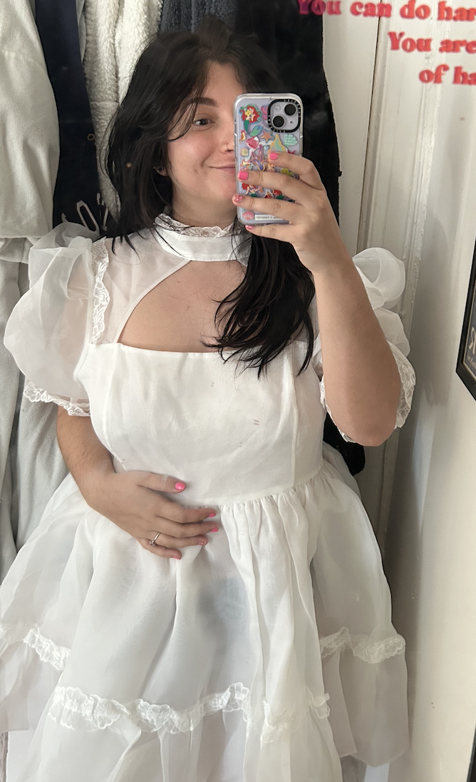 Person taking a mirror selfie, wearing a white vintage-inspired dress with ruffle details, holding a phone