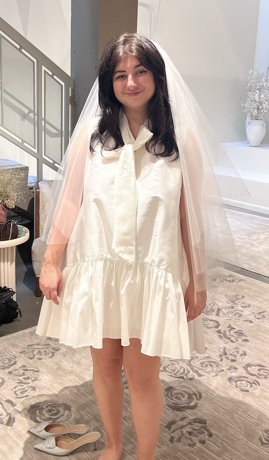 Person in a white short dress with a ruffled hem and a sheer veil, standing indoors