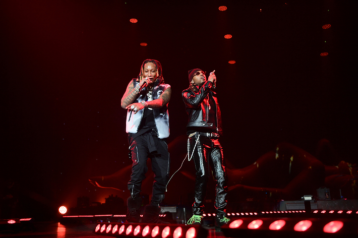 Two performers on stage, one in a black vest and jeans, the other in a red and black outfit, both singing into microphones