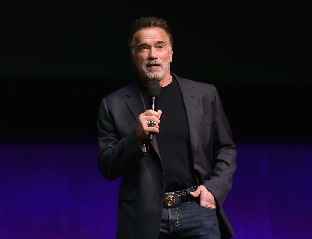 Arnold Schwarzenegger in a black jacket speaks into a microphone onstage