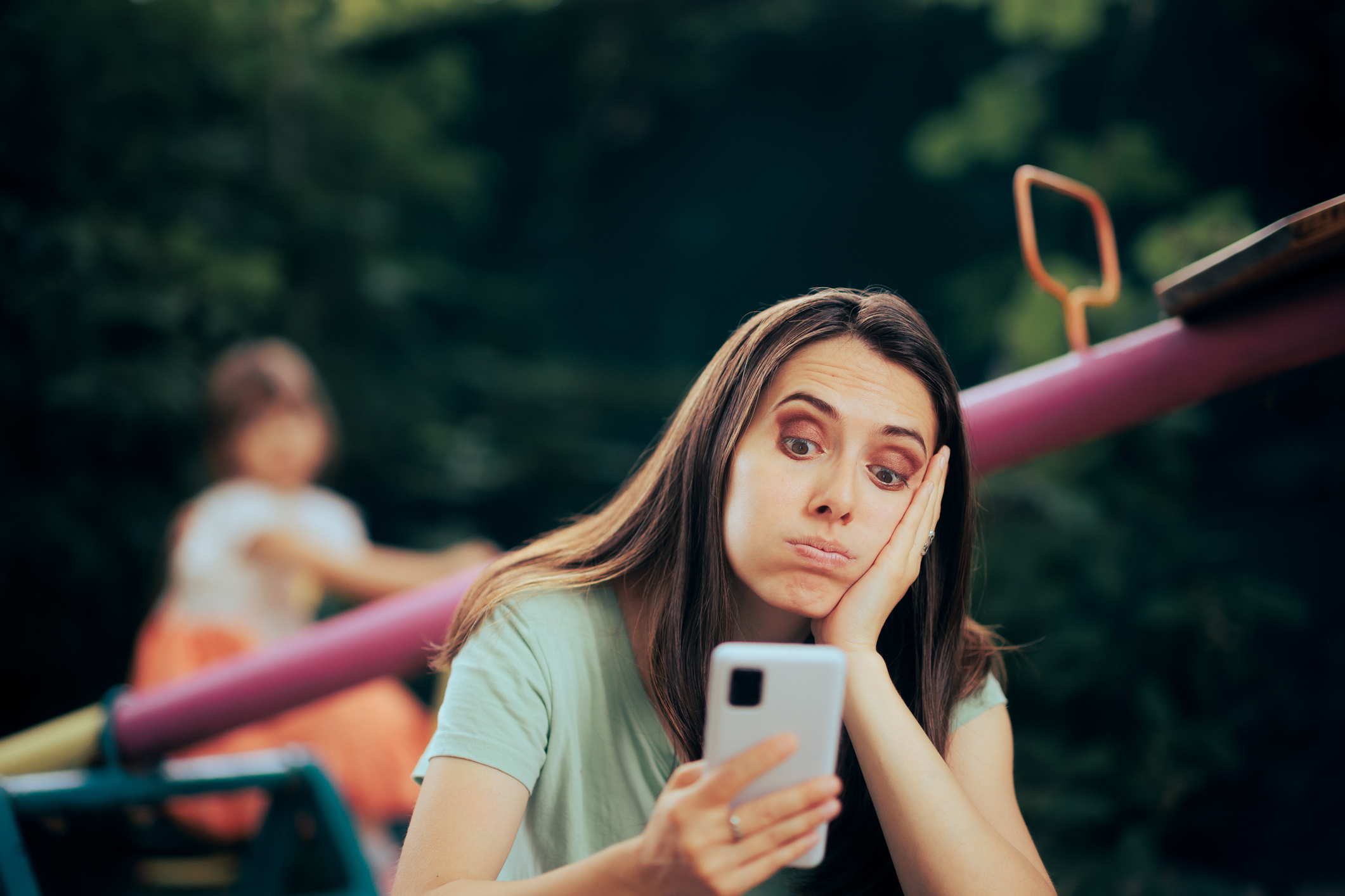 Woman looking at phone with bored expression, child in background on swing. They are outdoors, likely at a park