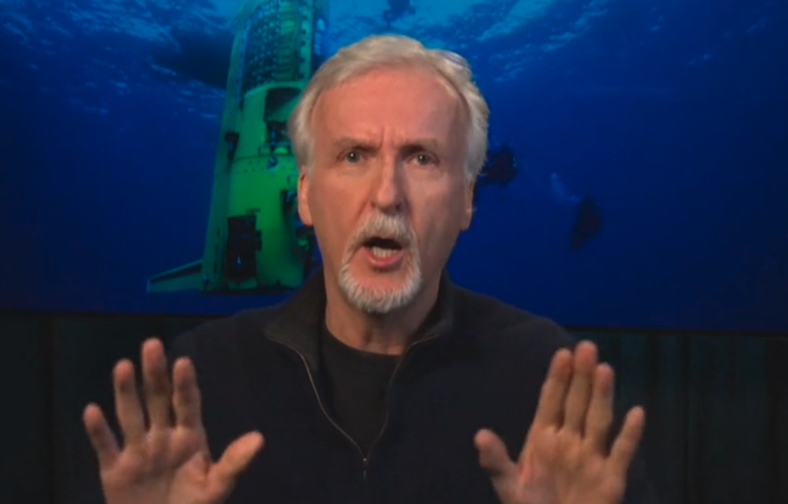 James Cameron with hands raised, speaking, with a submarine and divers in the background