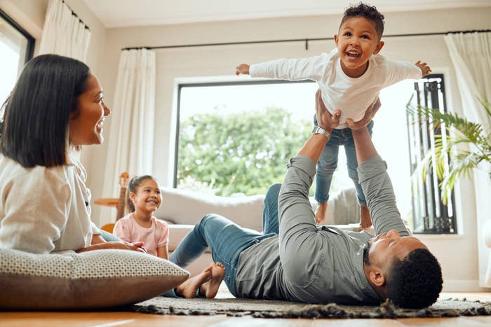 A family playing at home, with a parent lifting a laughing toddler in the air while another child and parent watch